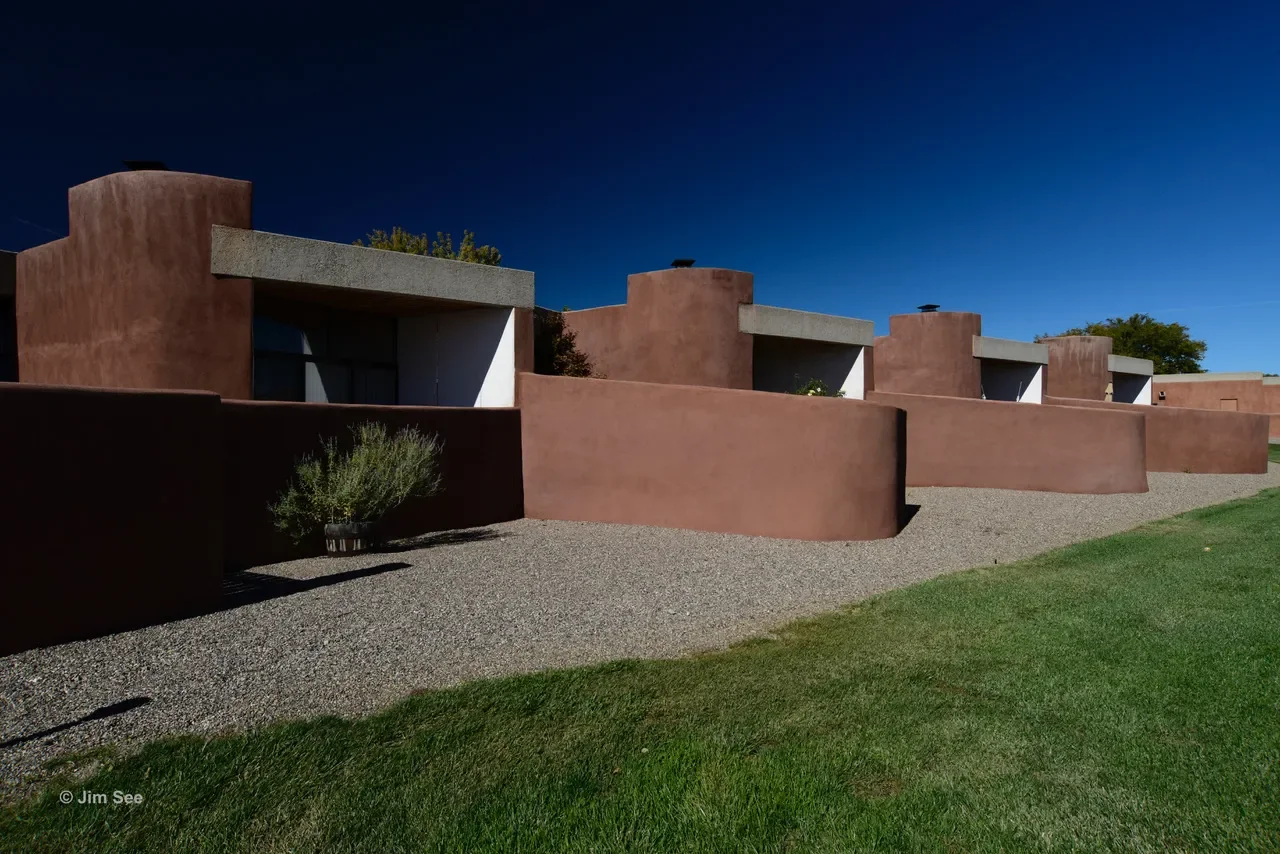 A row of modern, adobe-style houses with flat roofs and stucco exteriors, set against a deep blue sky. The scene includes gravel pathways, a small shrub, and a patch of grass.