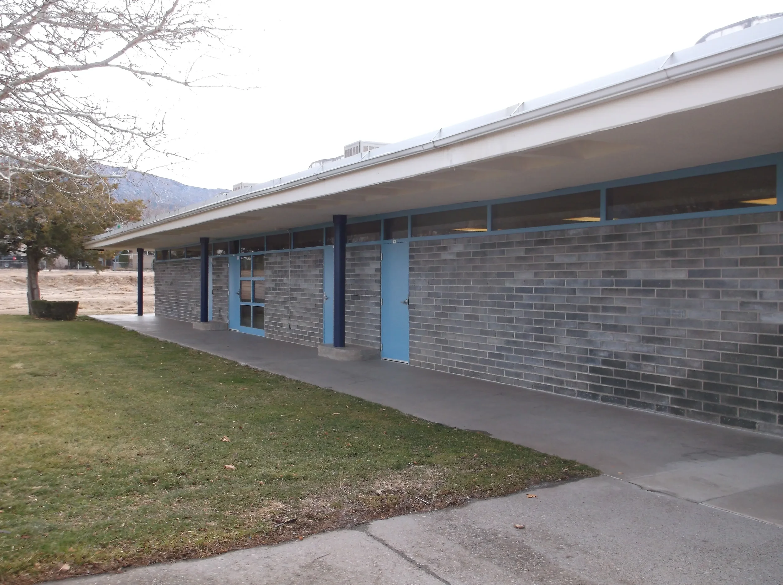 A brick building with blue accents and large glass windows, surrounded by grass and trees, with mountains visible in the background.