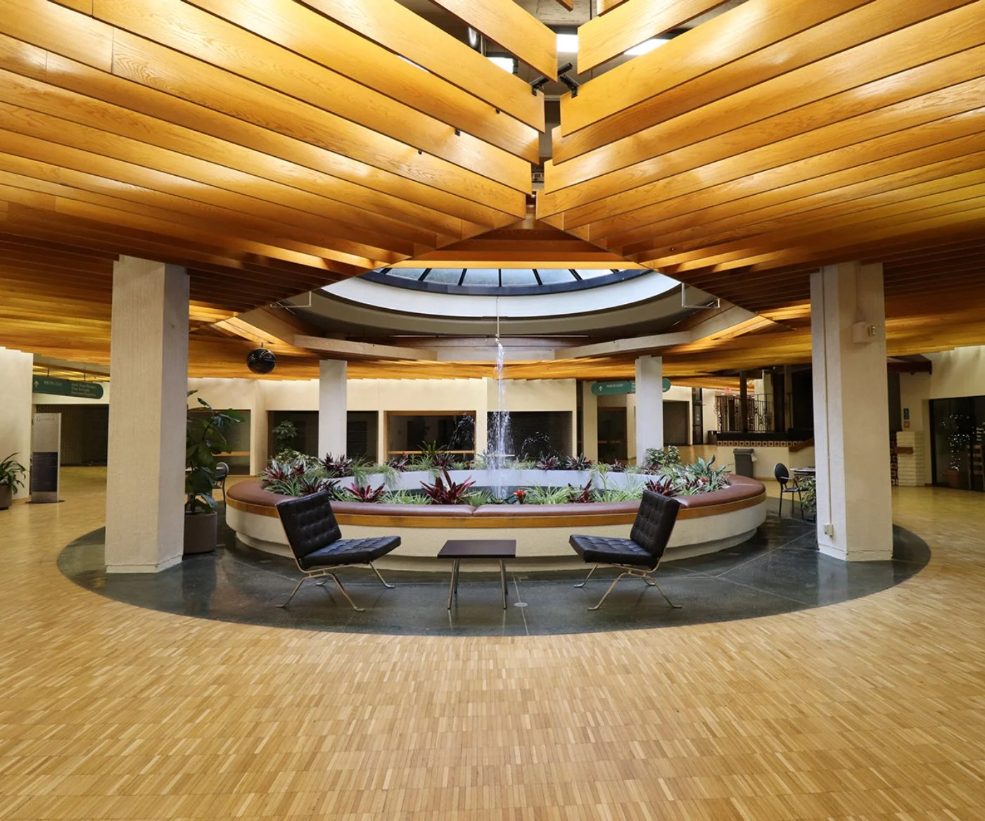 Interior view of a spacious building with a central circular water fountain surrounded by plants, chairs, and tables. The ceiling is made of wooden panels with a skylight overhead.