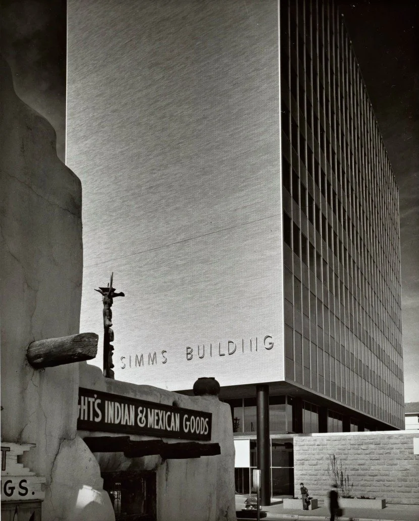 Black and white photo of a modern rectangular office building labeled 'Simms Building' with a rough-textured exterior and numerous windows. In the foreground, a traditional adobe-style sign for a shop selling Indian and Mexican goods features a wooden cross and a small sculpture of a crucifix.