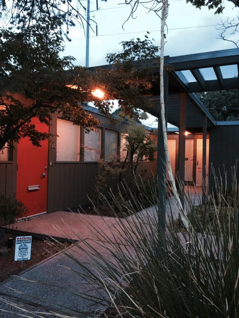 Exterior view of a modern building with a red door, large windows with closed blinds, and an entryway with a small sidewalk. There are trees, plants, and a warning sign on the ground near the sidewalk that reads "Warning: Armed Response".