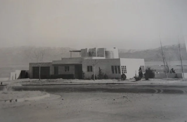 Black and white photograph of a modern, mid-century style house situated in a barren landscape with distant hills, a few trees, utility poles, and a curved road in the foreground.