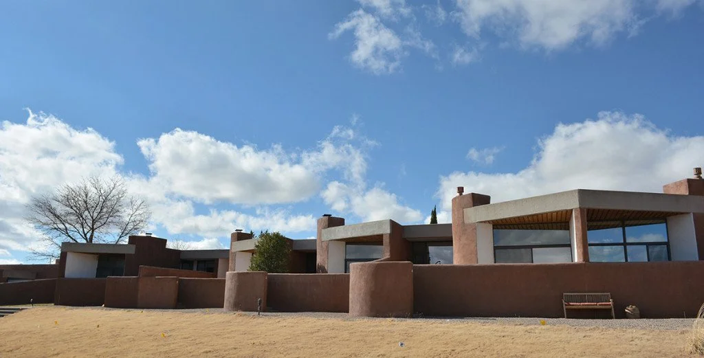 A modern house with a flat roof, large windows, and a stucco exterior in earthy tones, set against a partly cloudy blue sky with a leafless tree nearby.
