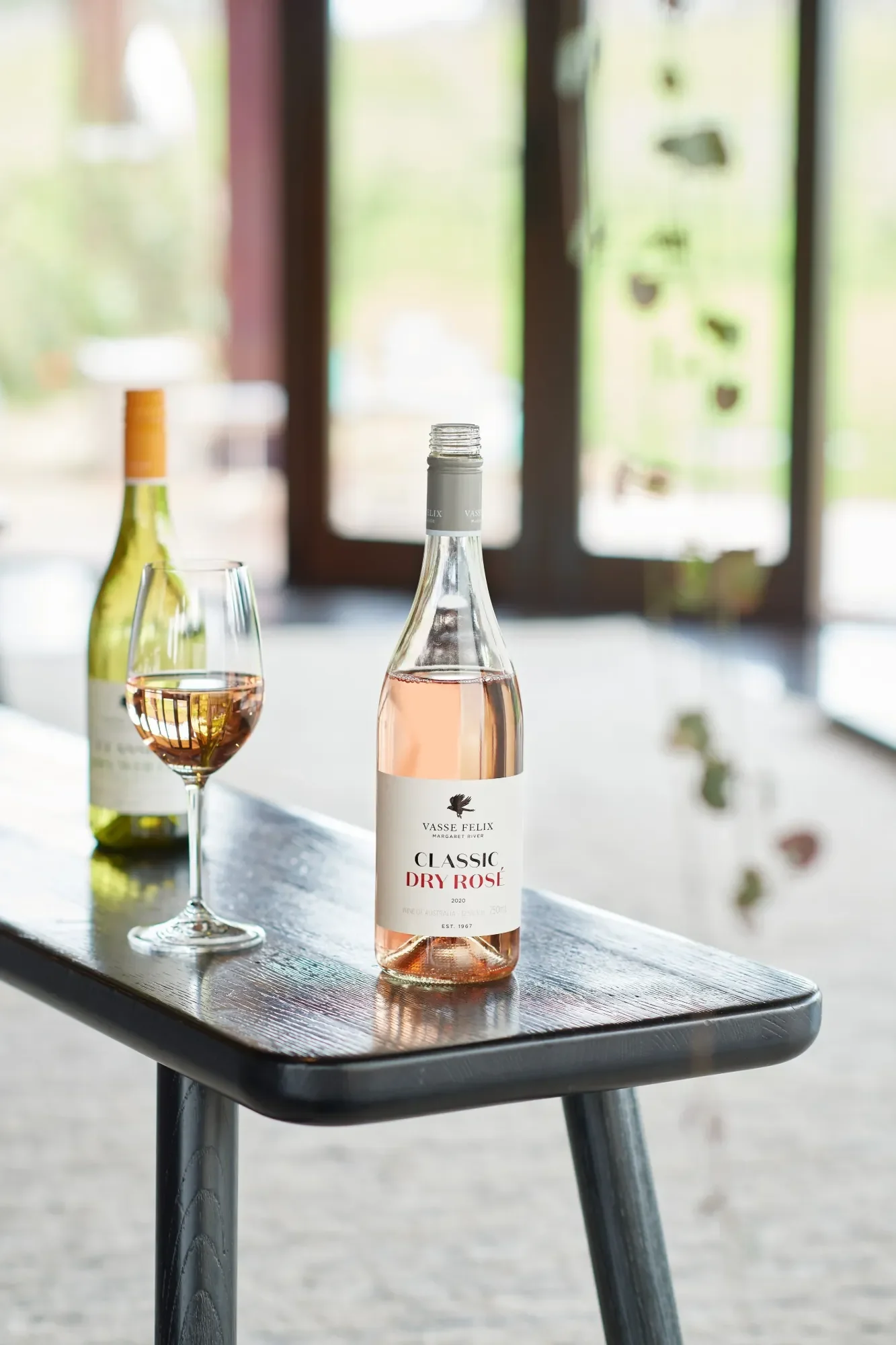 A bottle of Vasse Felix Classic Dry Rosé wine and a glass filled with rosé, placed on a dark wooden table with a blurred background of a window and hanging decorations.