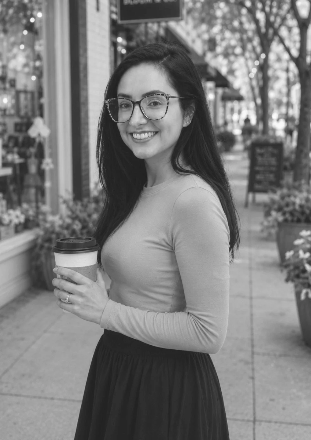 Black-and-white photo of a smiling woman with long dark hair and glasses, wearing a fitted long-sleeve top and skirt, holding a coffee cup while standing on a charming downtown sidewalk with storefronts, flowers, and trees in the background.