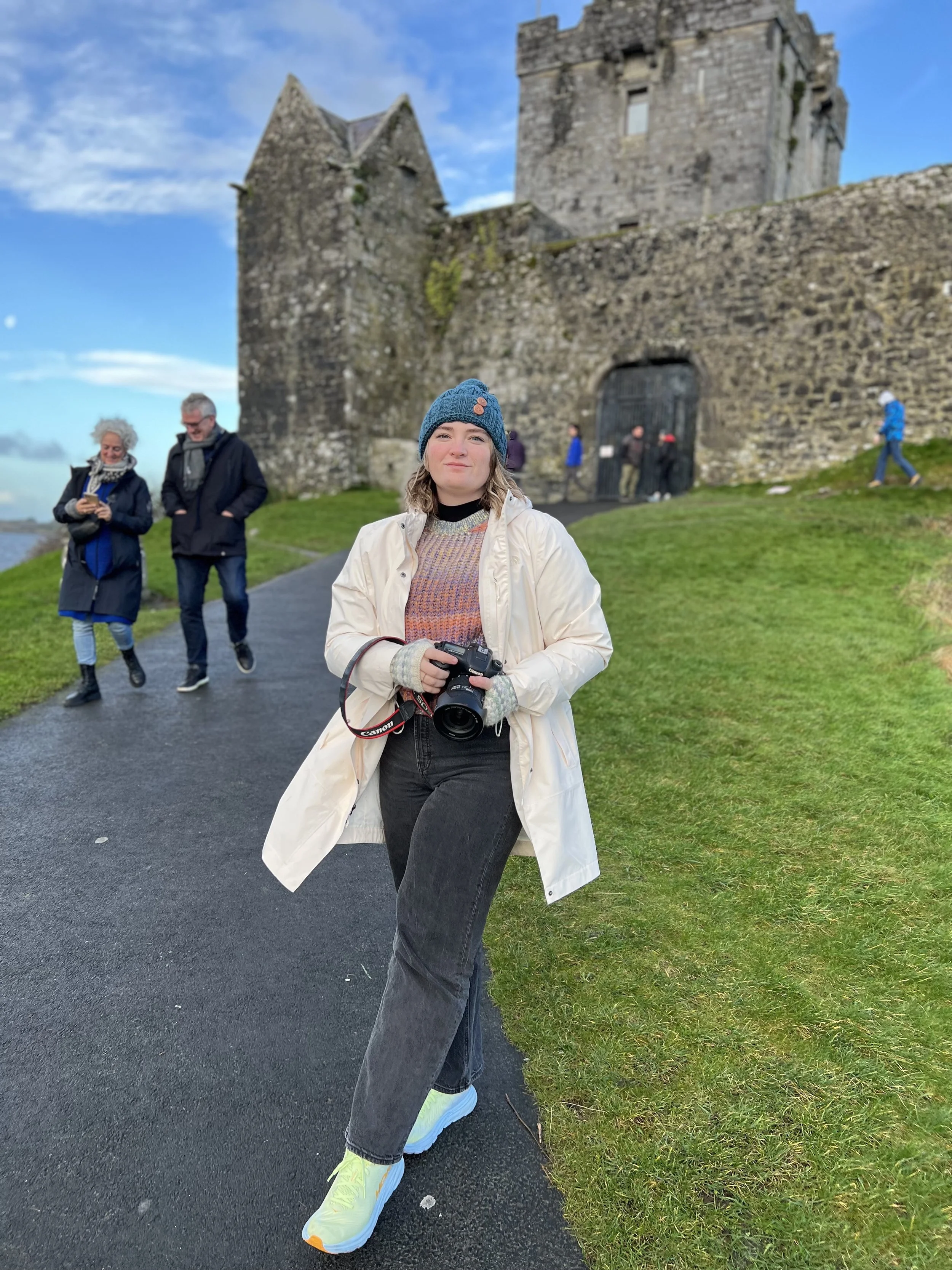 A woman standing on a paved pathway in front of an old stone castle, holding a camera, wearing a beanie, light-colored coat, black pants, and light-colored sneakers, with other people walking and exploring the castle in the background.