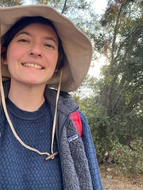A woman outdoors wearing a wide-brimmed hat, a blue sweater, a fleece vest, and a red backpack, smiling at the camera with trees in the background.