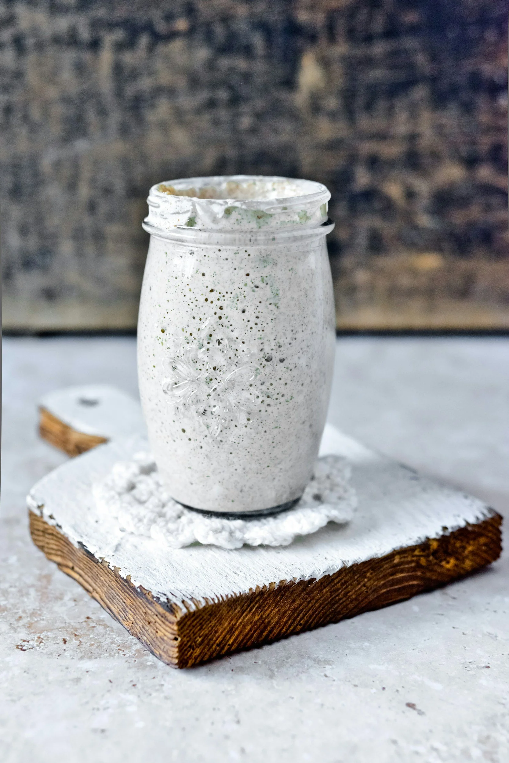 A glass jar with a textured, frosted appearance containing a creamy, speckled beverage, placed on a white cloth on a rustic wooden board with a dark, textured background.