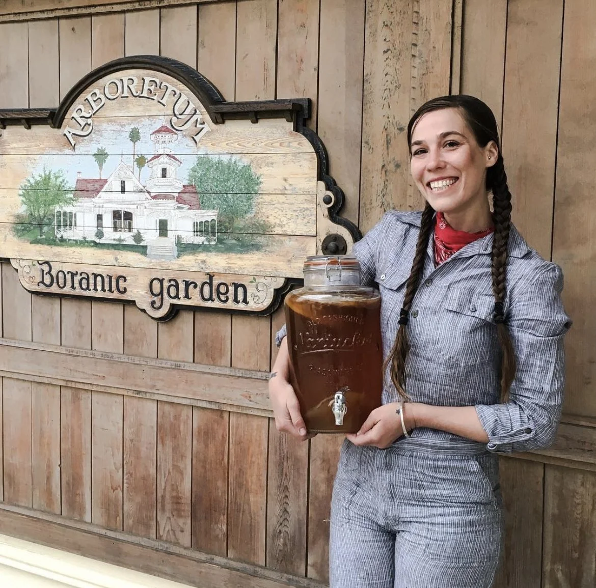 A woman smiling and holding a large jar with a tap, filled with a dark beverage, standing in front of a wooden wall with a sign that reads 'ABBREITUNG Botanic garden' and features an illustration of a large house surrounded by trees.
