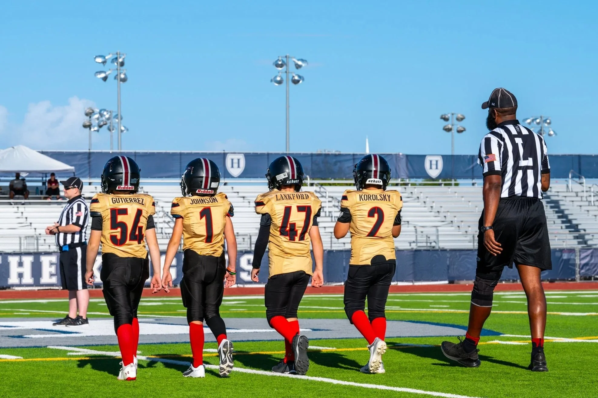 American football team walking on the field with four players wearing gold jerseys and black helmets, alongside a referee, on a bright sunny day with stadium lights and empty bleachers in the background.