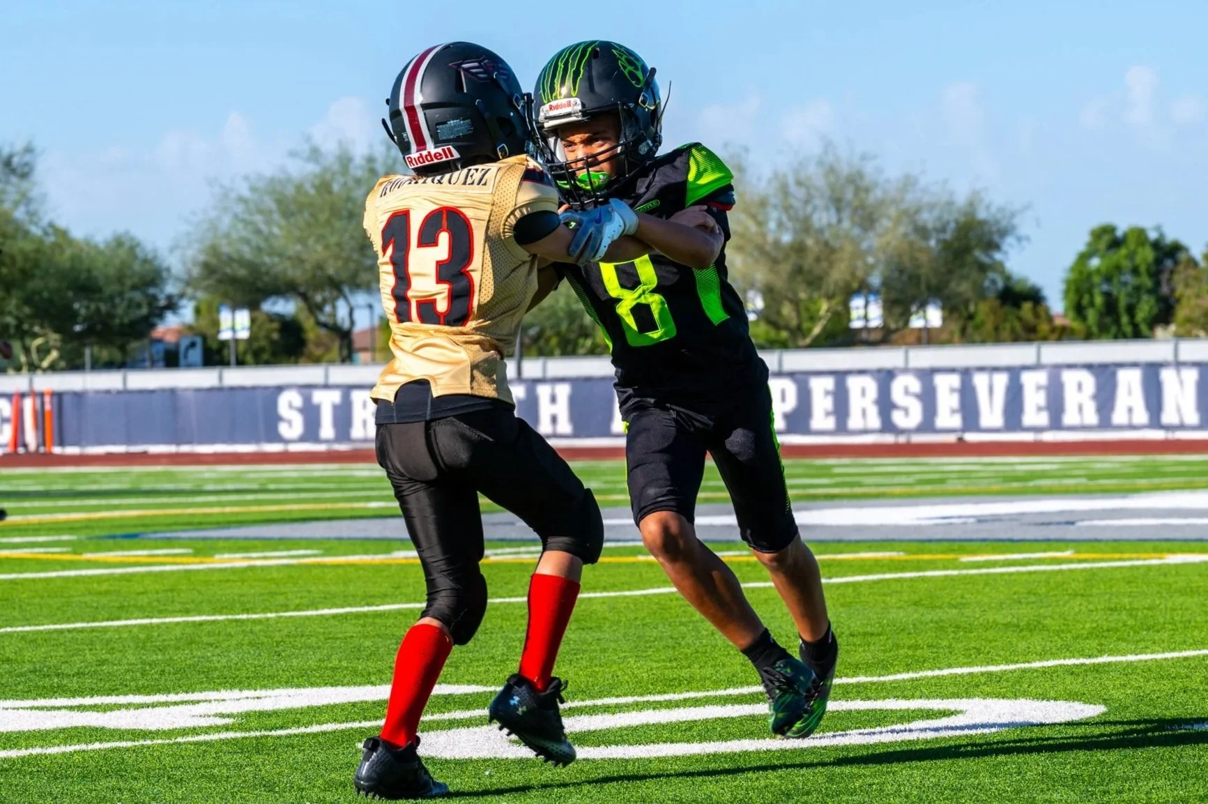 Two young football players are engaged in a clash on a football field, with one wearing a gold jersey with the number 13 and the other in a black and green jersey with the number 8. A referee and other players are visible in the background on the sun