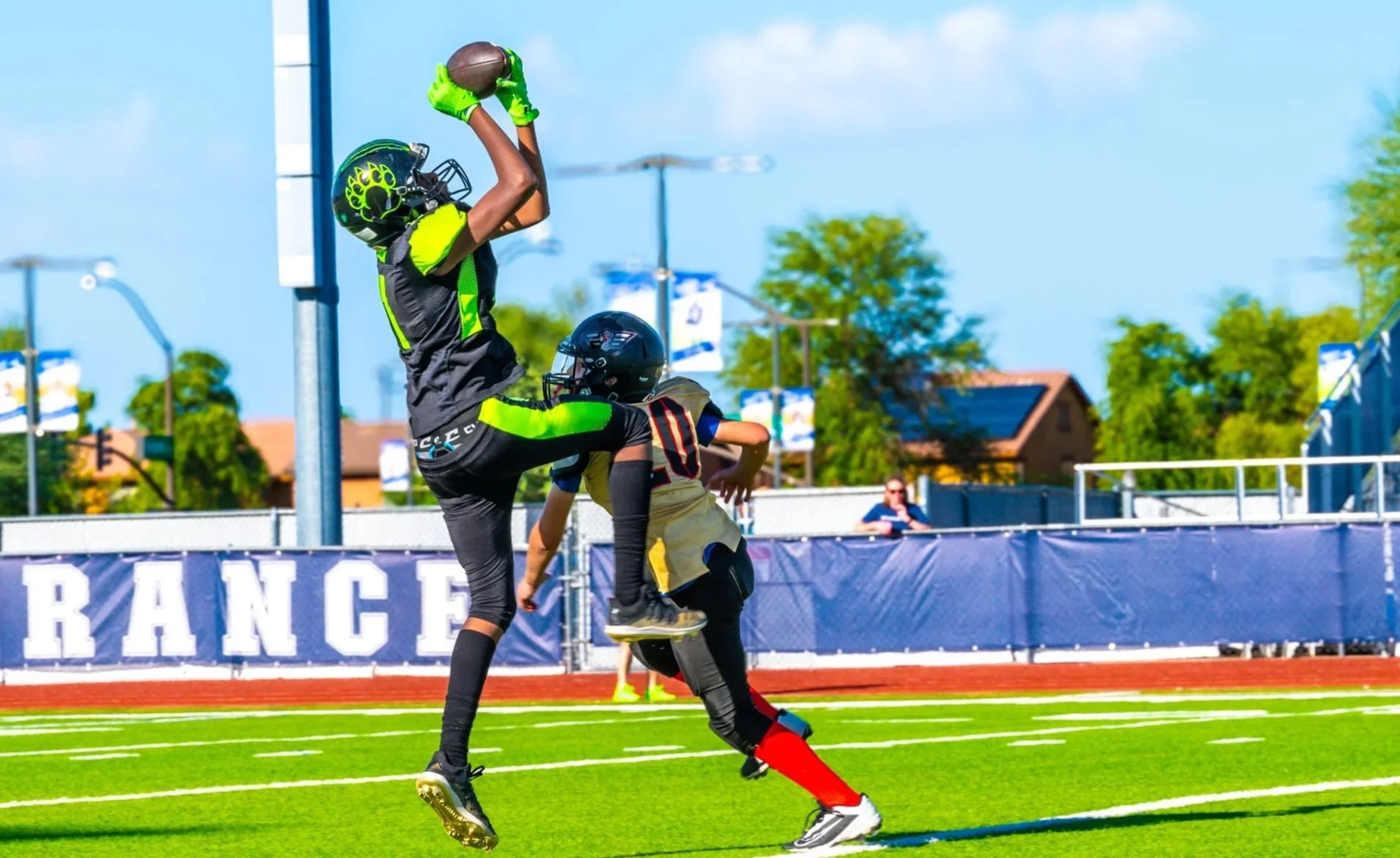 Two young football players in helmets and uniforms compete for a football in the end zone, with one jumping to catch it while the other defends. The game takes place on a green field with stadium seating and a banner reading 'PERSEVERANCE' in the bac
