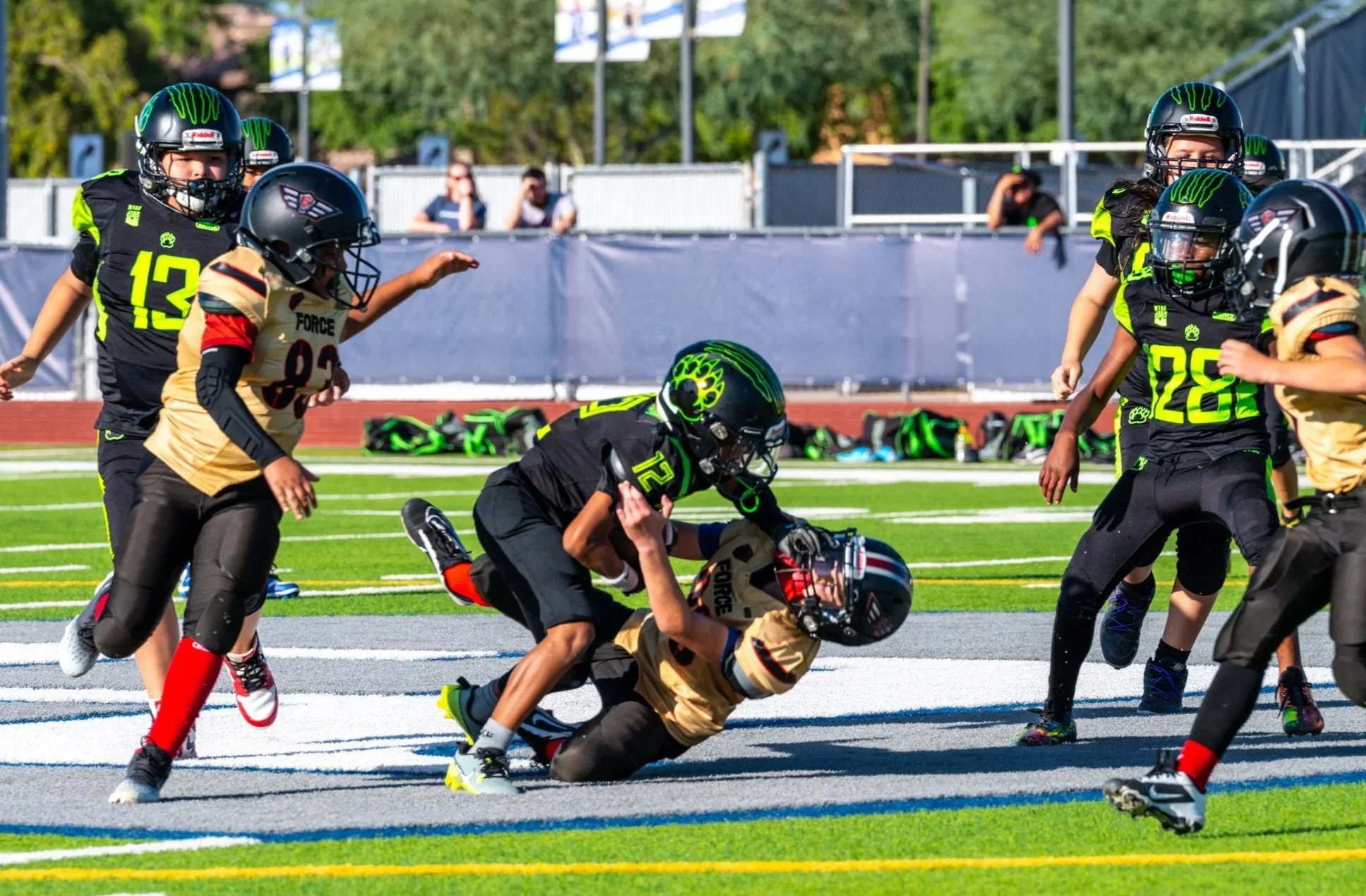 Youth football game with players in black and gold uniforms on a bright green field, some tackling and running with the ball, on a sunny day.