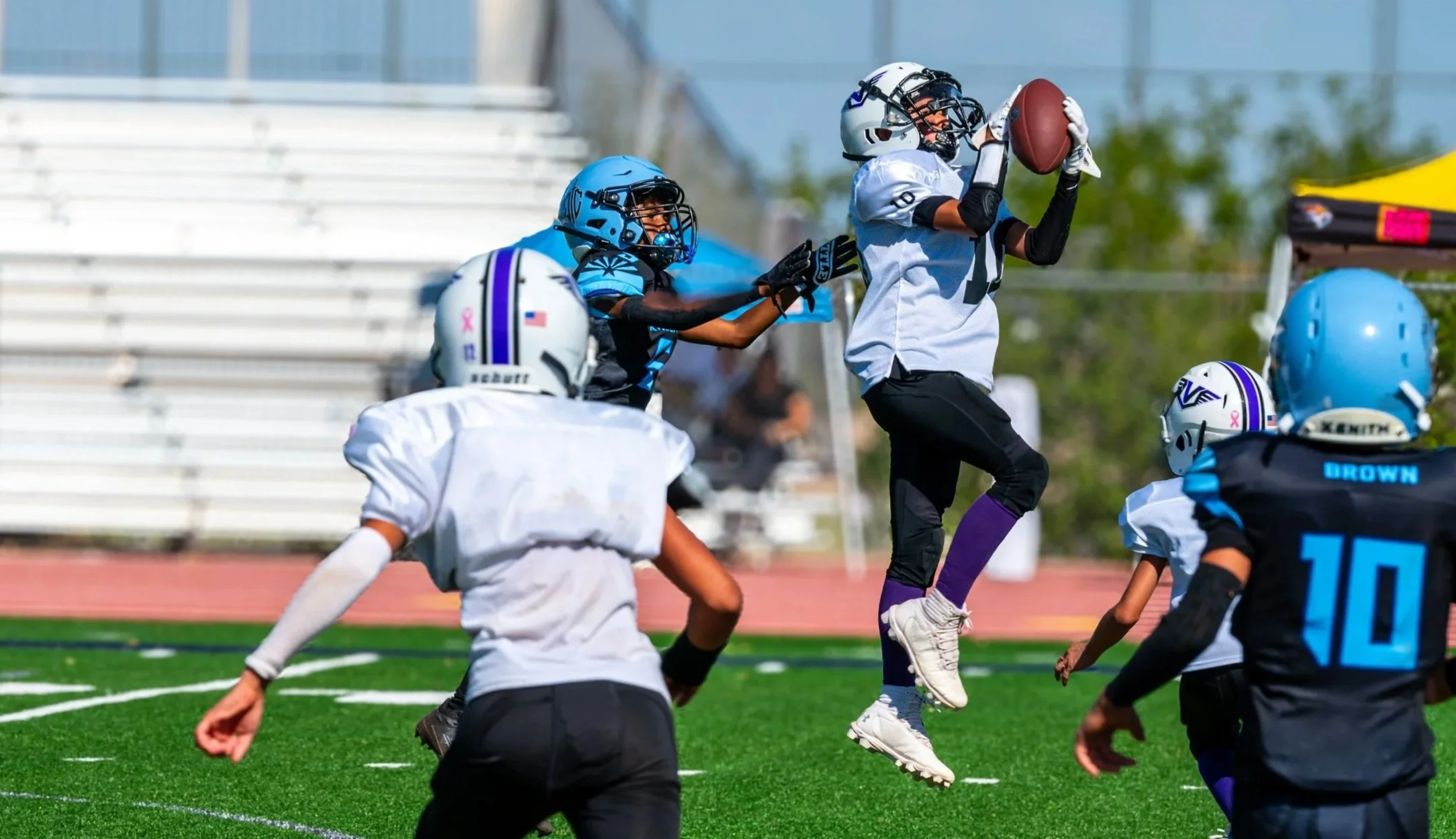 Young football players in helmet and pads during a game on a sunny day, with one player jumping to catch the football while others try to block.