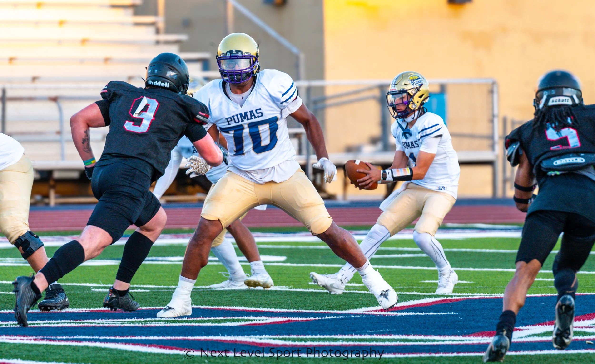High school football game with players in action on the field, one team in black and the other in white with gold helmets.