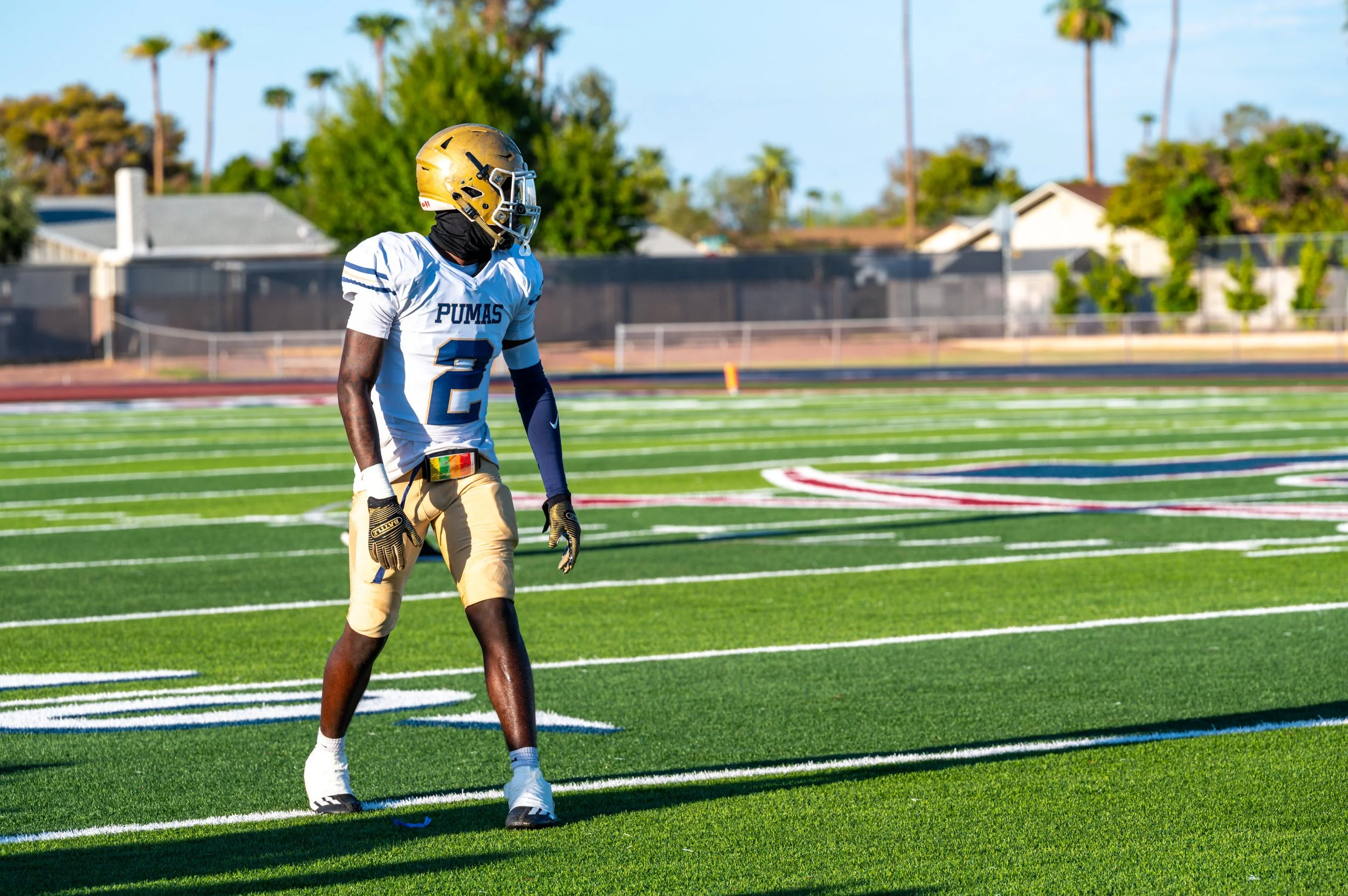 A football player wearing a white jersey with the number 2 and 'Pumas' on it, gold helmet, and gold shorts, standing on a football field.