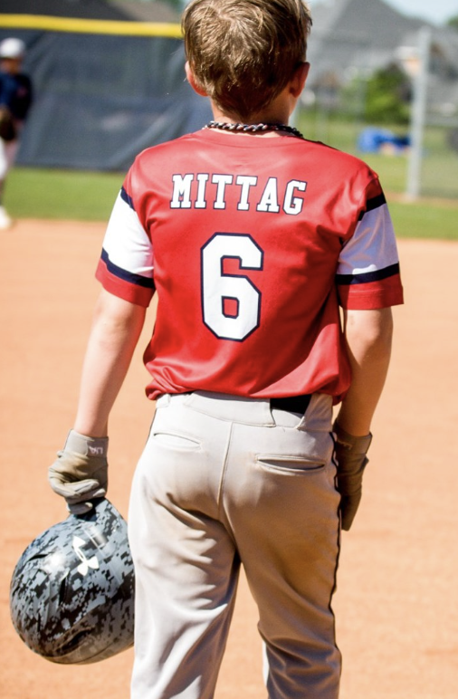 Back view of a young baseball player wearing a red jersey with 'MITTAG 6' on the back, holding a camouflage helmet in his right hand on a baseball field.