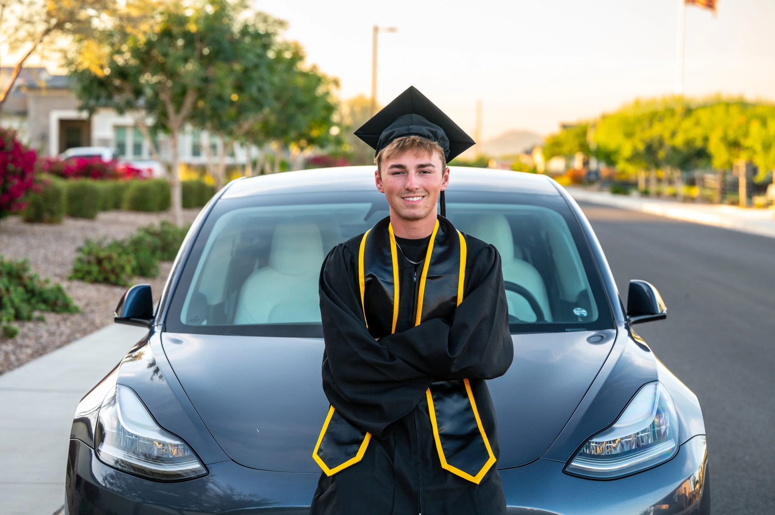 A young man in a black graduation gown and cap with yellow accents, standing in front of a car on a suburban street, smiling at the camera