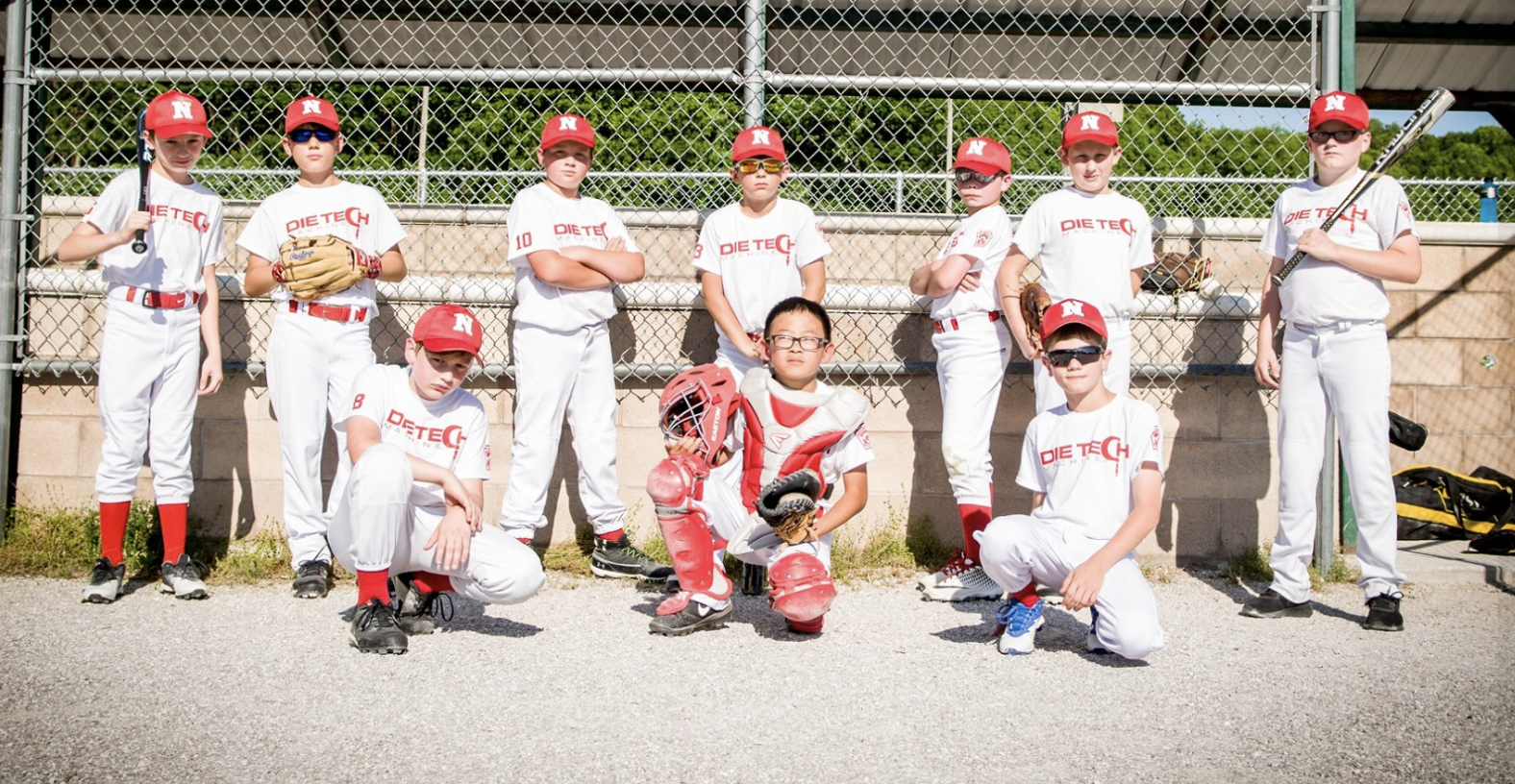 Youth baseball team posing for a group photo on a baseball field. The players are in white uniforms with red caps and equipment, standing and kneeling in front of a chain-link fence.