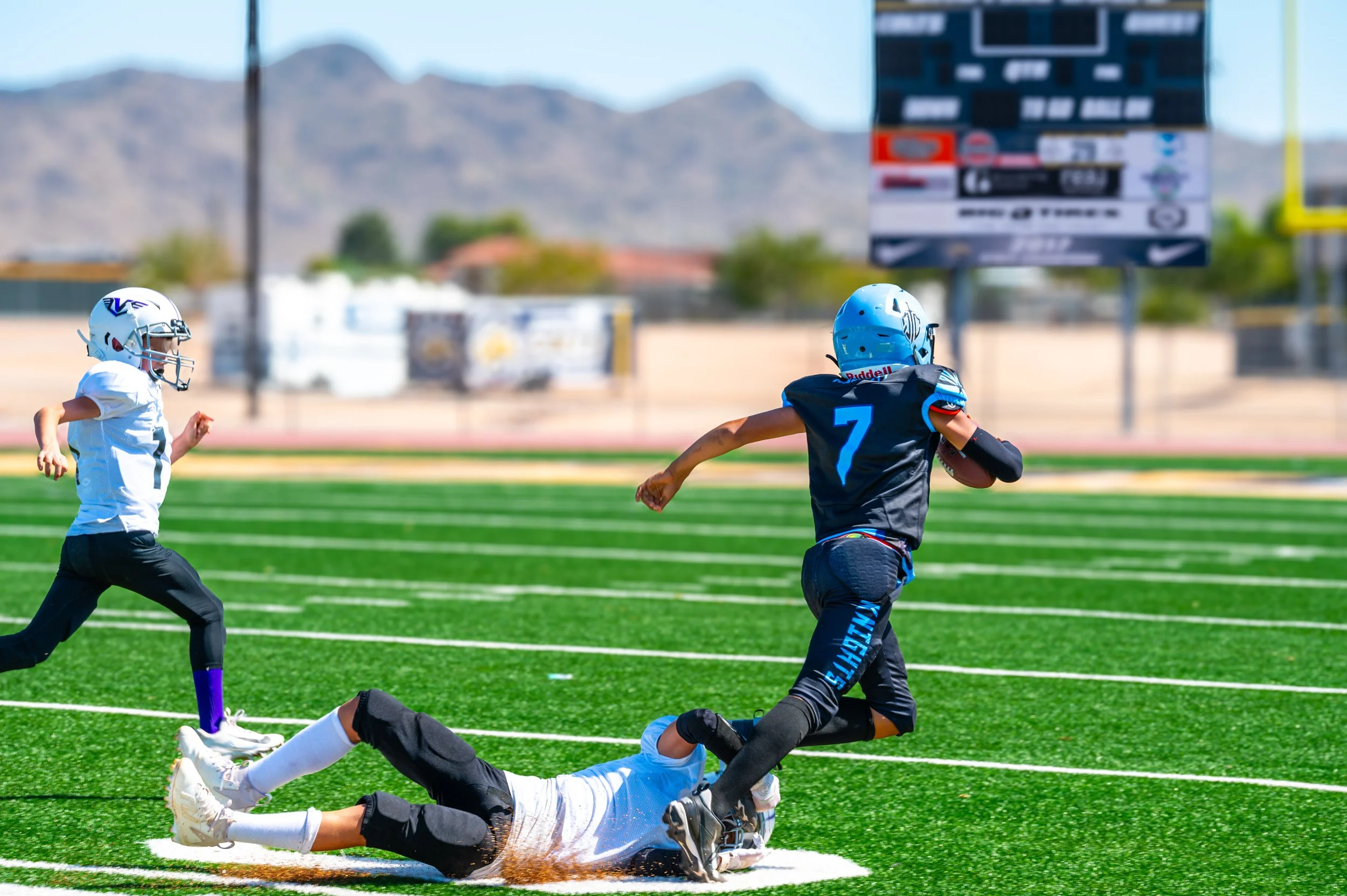 A young football player wearing a blue helmet, black jersey with the number 7, running with the football on a green field, while a player in a white uniform with a helmet and black pants is on the ground attempting to tackle him, with another player