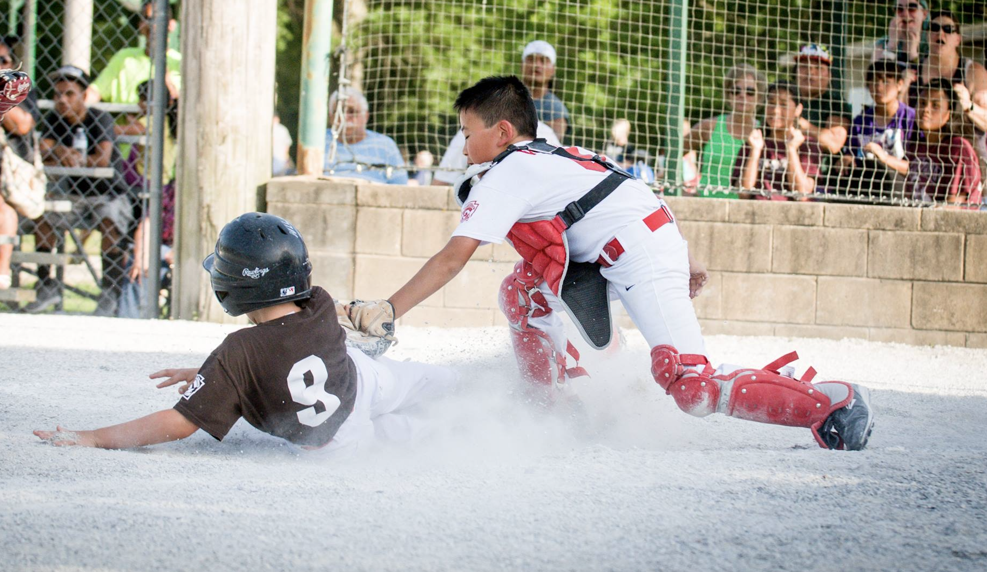 A baseball player in brown and white uniform sliding on the ground while being tagged by a catcher in white and red gear during a game, with spectators watching behind a fence.