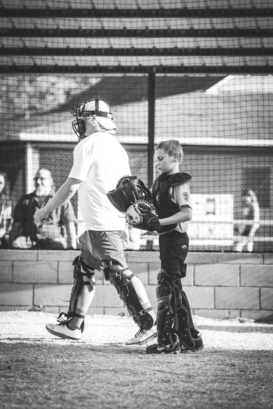 A young baseball catcher in gear standing on the field, with an adult in catcher's gear, in black and white, at a baseball field with a chain-link fence and spectators in the background.