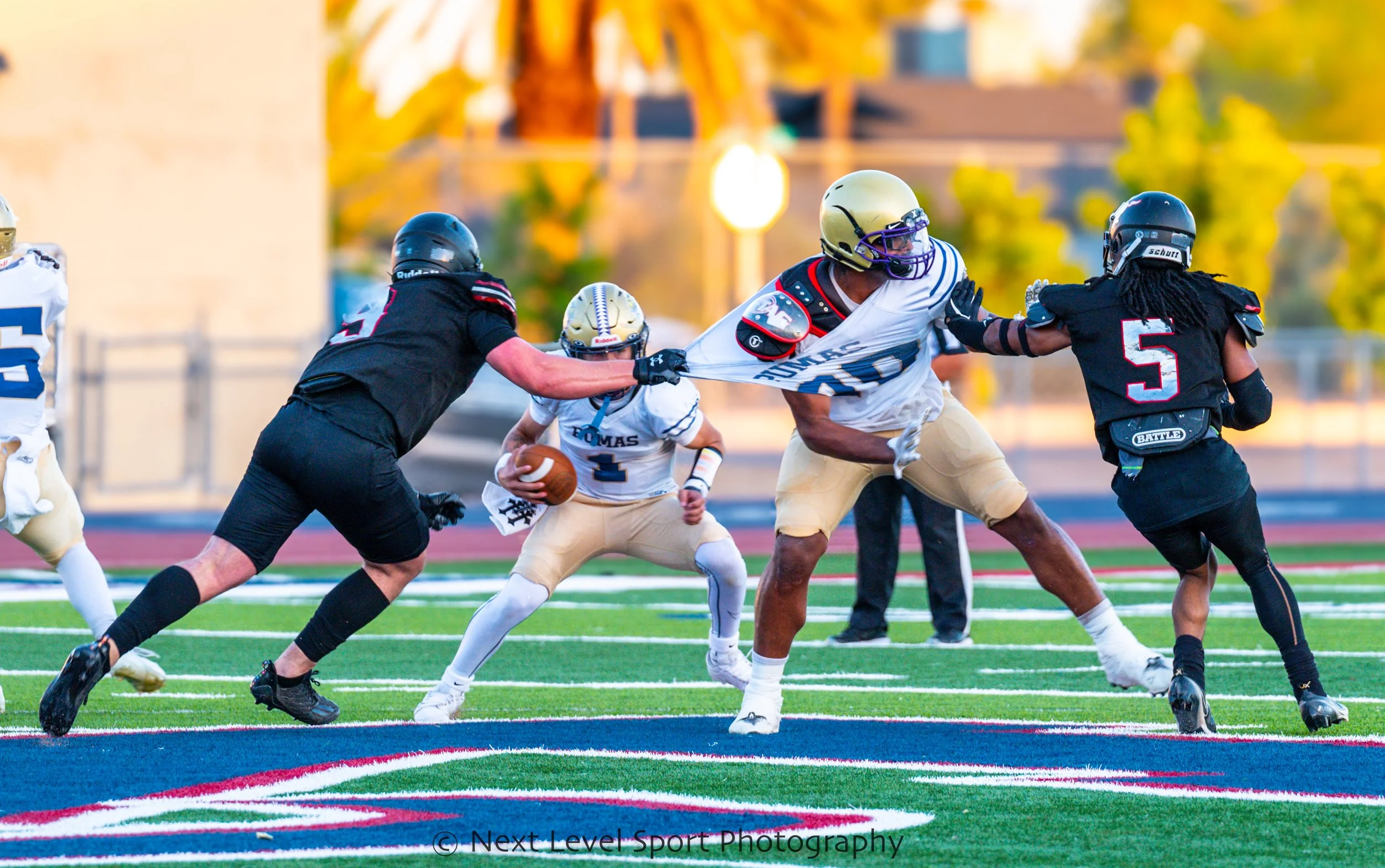 A youth American football game where a player in white and gold uniform runs with the football while players in black uniforms attempt to block or tackle him on a football field with vibrant green turf and colorful markings.