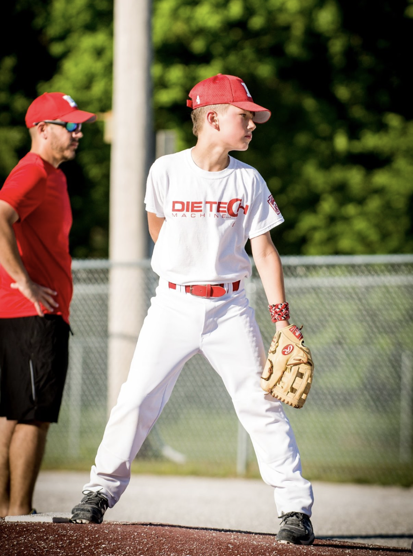 A young baseball player in a white uniform and red cap stands on the mound, wearing a glove, with an adult coach in a red shirt and cap nearby on a baseball field.