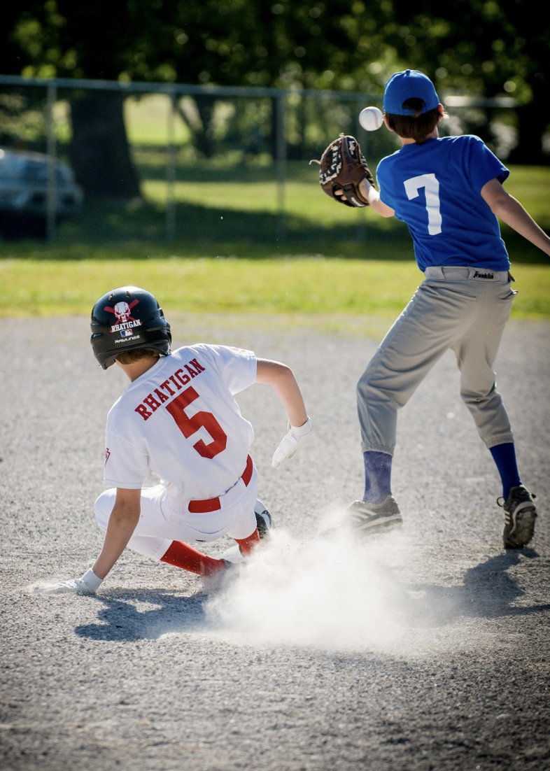 Children playing baseball on a field, wearing helmets and uniforms, with one sliding into a base and another catching a ball.