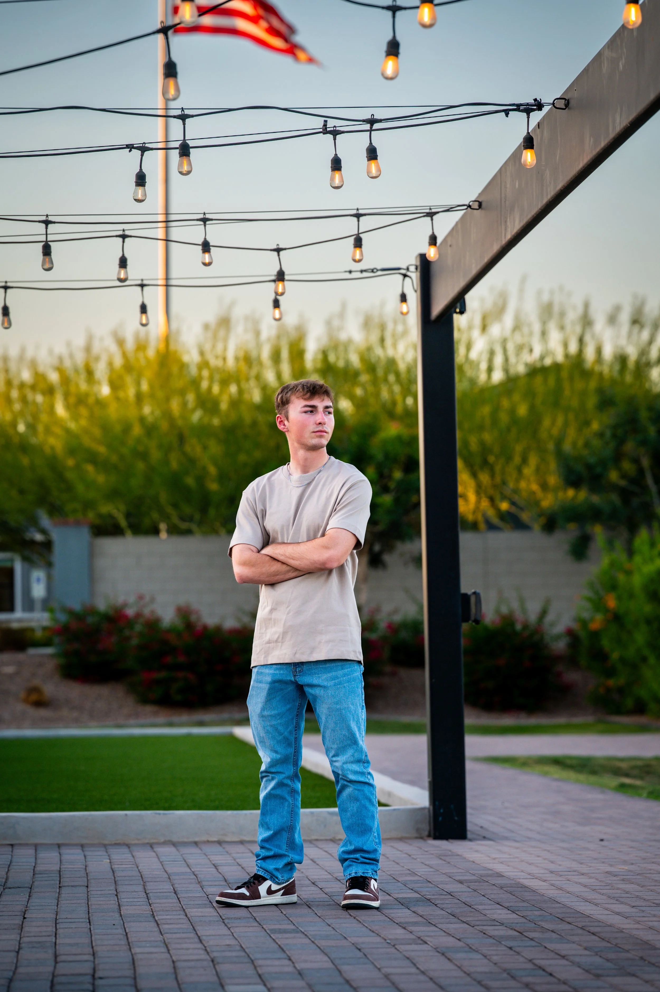 Young man standing with arms crossed outdoors during evening, under string lights and with an American flag in the background.