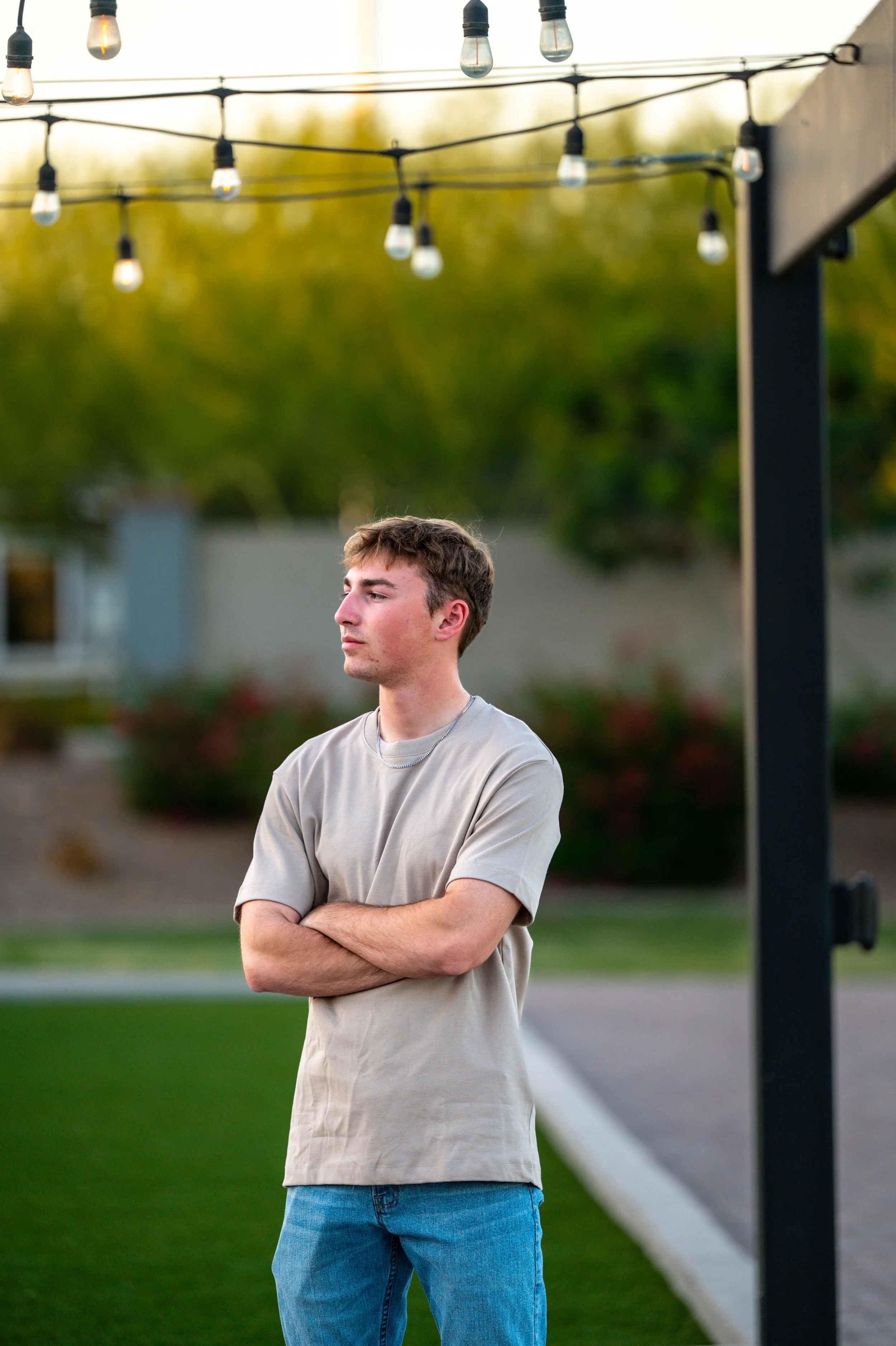 Young man standing outdoors with cross arms under string lights, looking to the side during sunset.