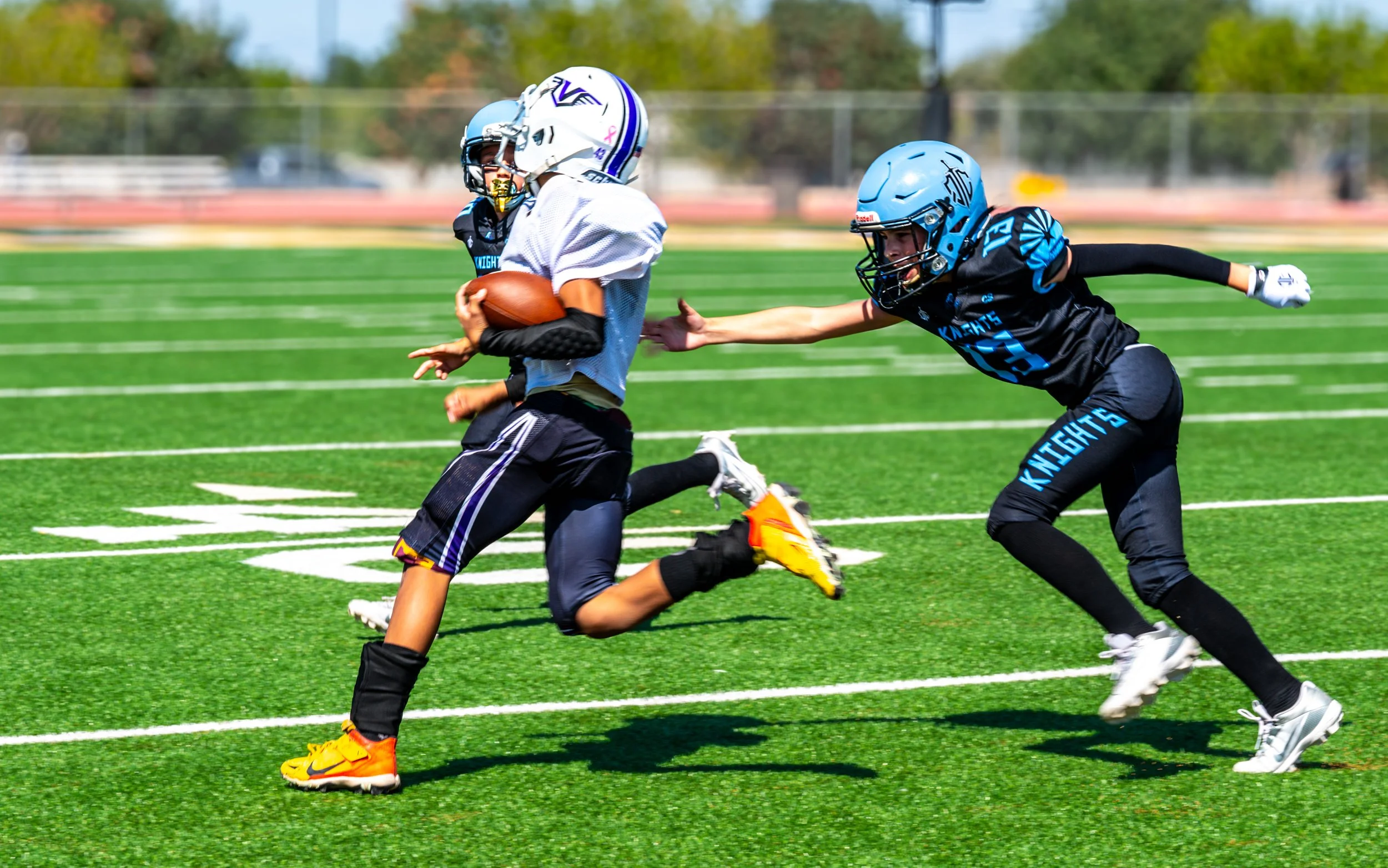 A youth football game on a green field with white yard lines. A player in a white jersey is running with the ball while a player in a black jersey with blue accents reaches out to try to tackle him.