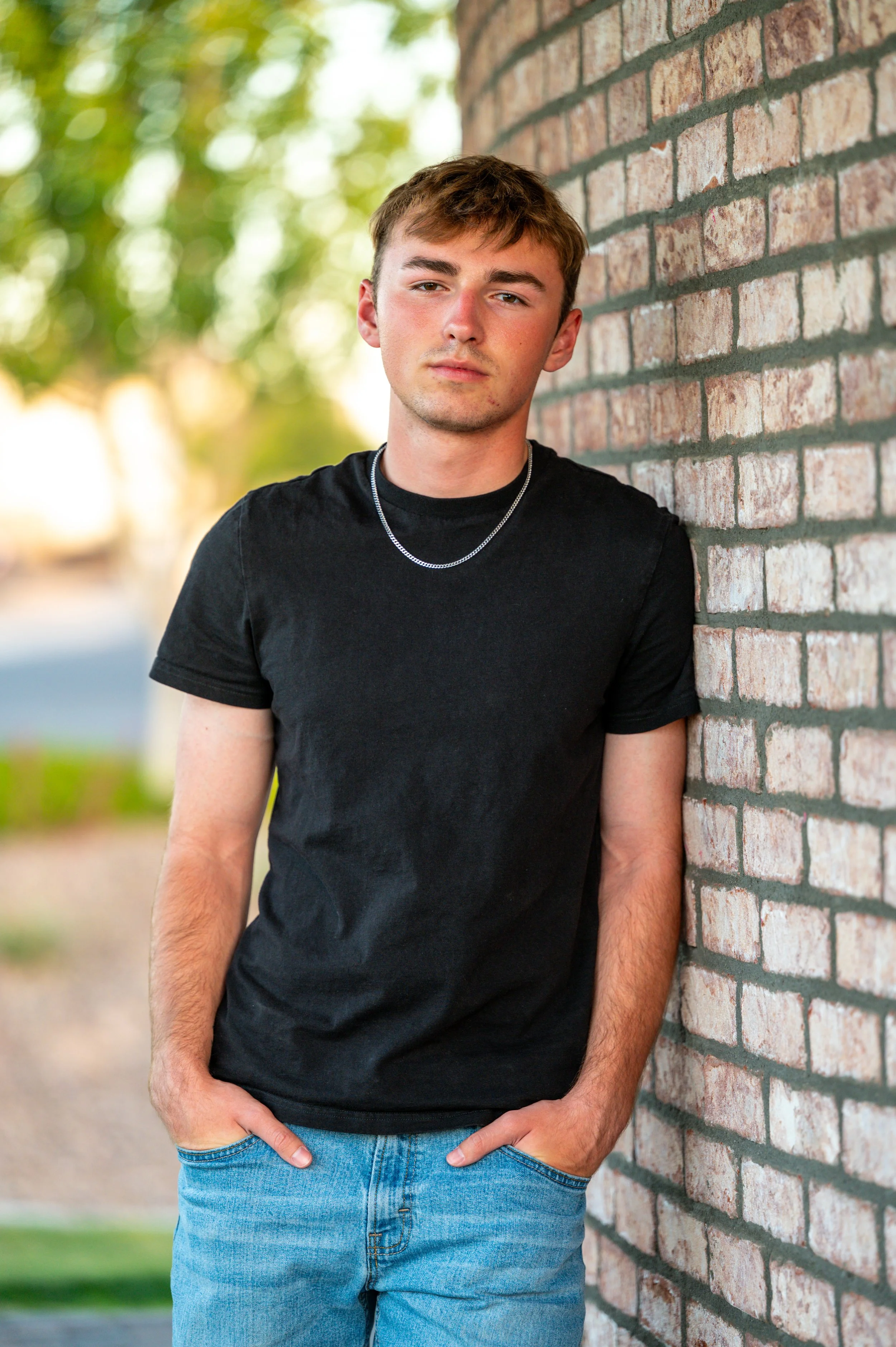 A young man with short light brown hair wearing a black t-shirt and light blue jeans, standing outdoors against a brick wall with a blurred background of trees and greenery.