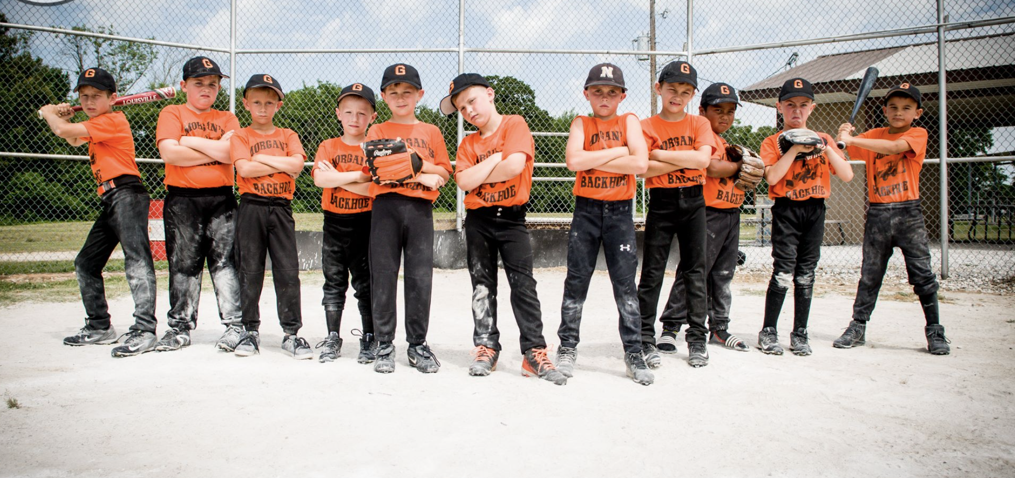Young boys in orange team shirts and black pants standing on a baseball field, some holding baseball bats and gloves, posing for a team photo.