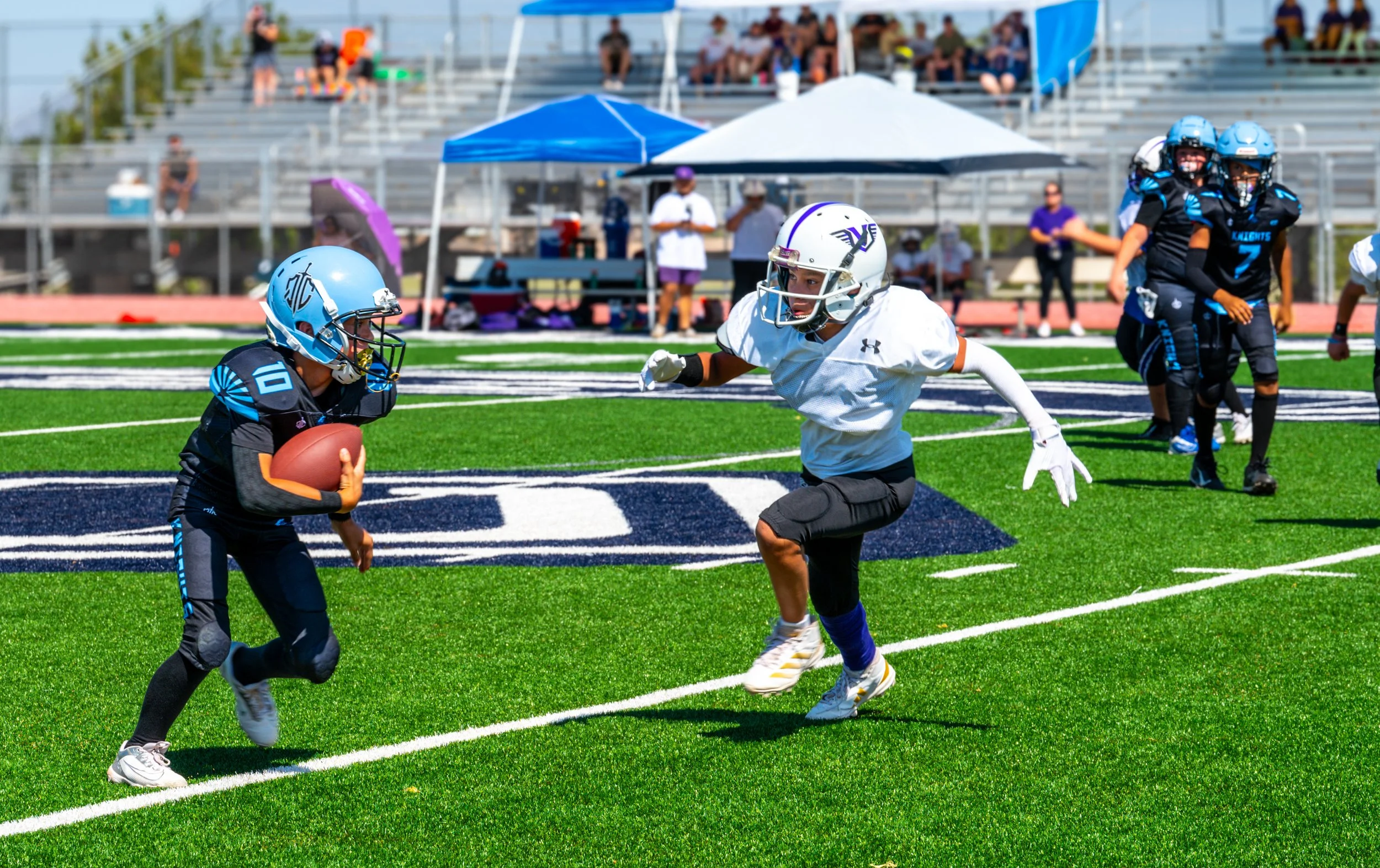 A youth football game on a green field with players in blue and black uniforms. A young player with a black uniform and helmet is running with the football, while a player in a white uniform and helmet is rushing toward him. Spectators sit in bleache