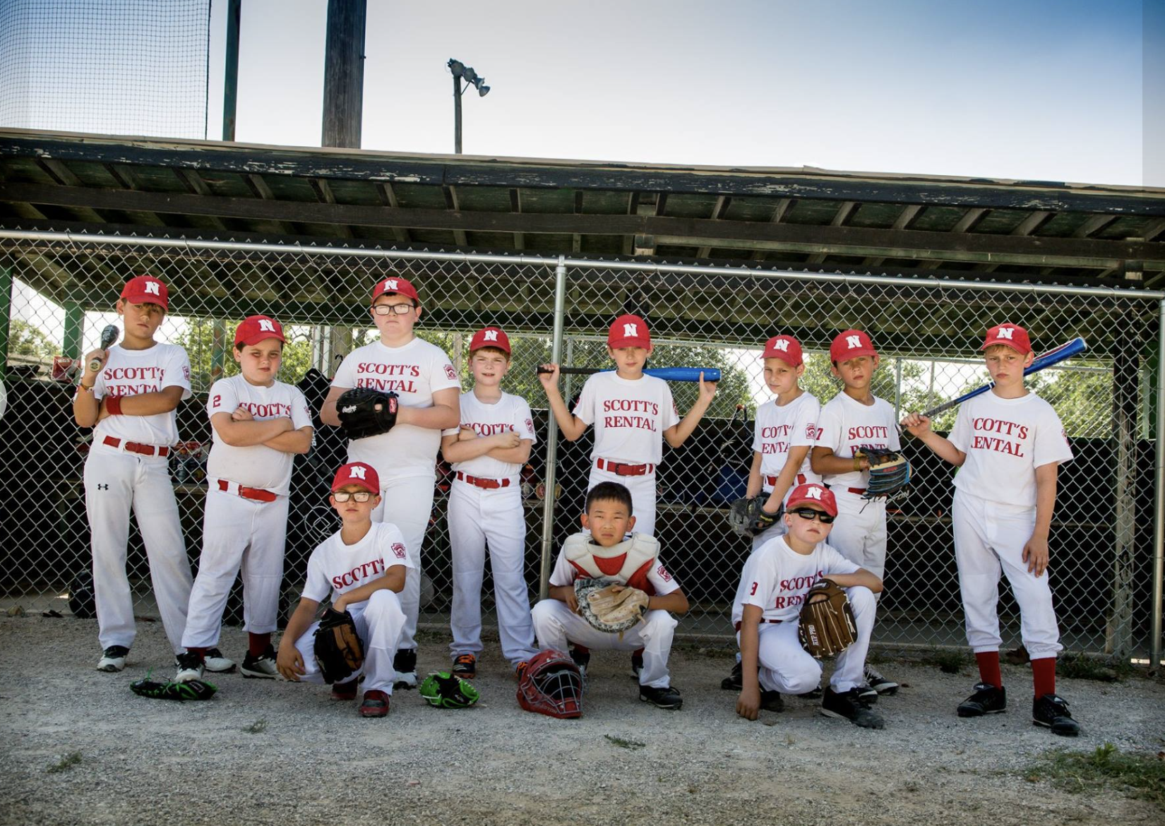 Youth baseball team in uniform posing for a team photo in front of a chain-link fence dugout, some players holding bats, others with gloves, wearing red caps with an 'N', and white jerseys with red lettering. Two players are kneeling in front, one wi