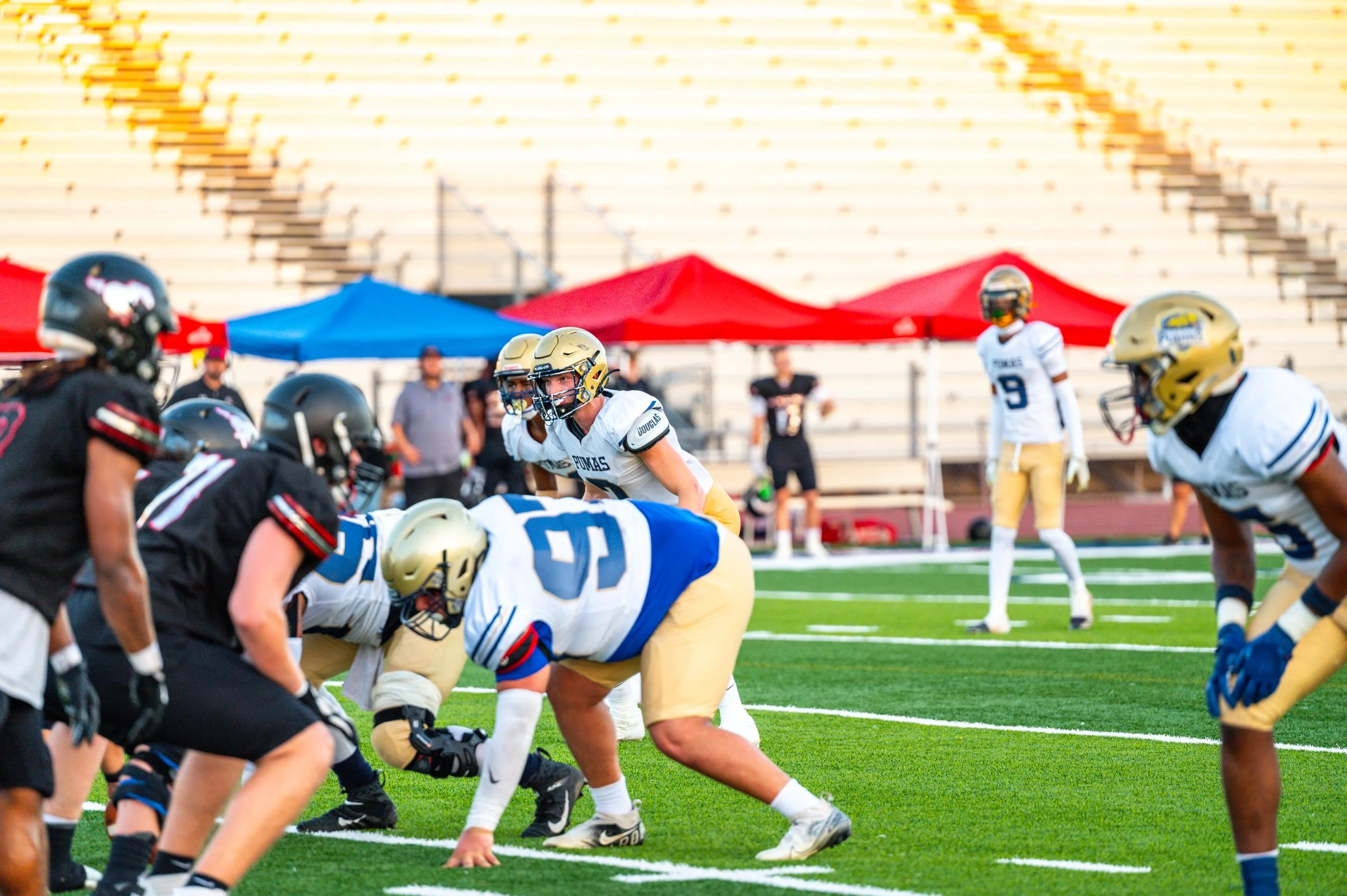 American football players on the field during a game, with players in white and gold uniforms facing players in black uniforms, and red and blue tents in the background.