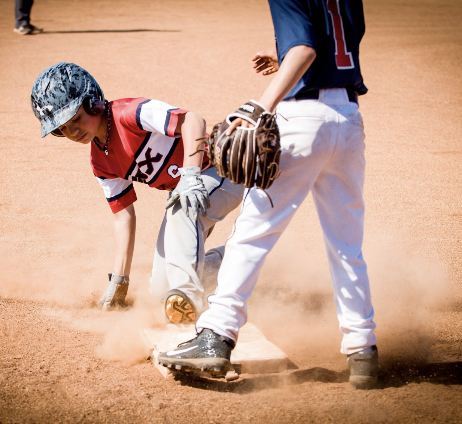A young baseball player in a red and white uniform is slipping on the base while an umpire stands nearby on the orange dirt field.