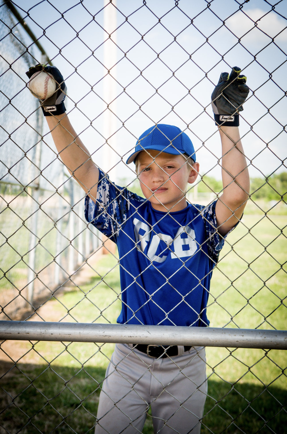 Young boy in a blue baseball uniform and cap standing behind a chain-link fence, holding a baseball in one arm and gripping the fence with both hands, on a baseball field during daytime.