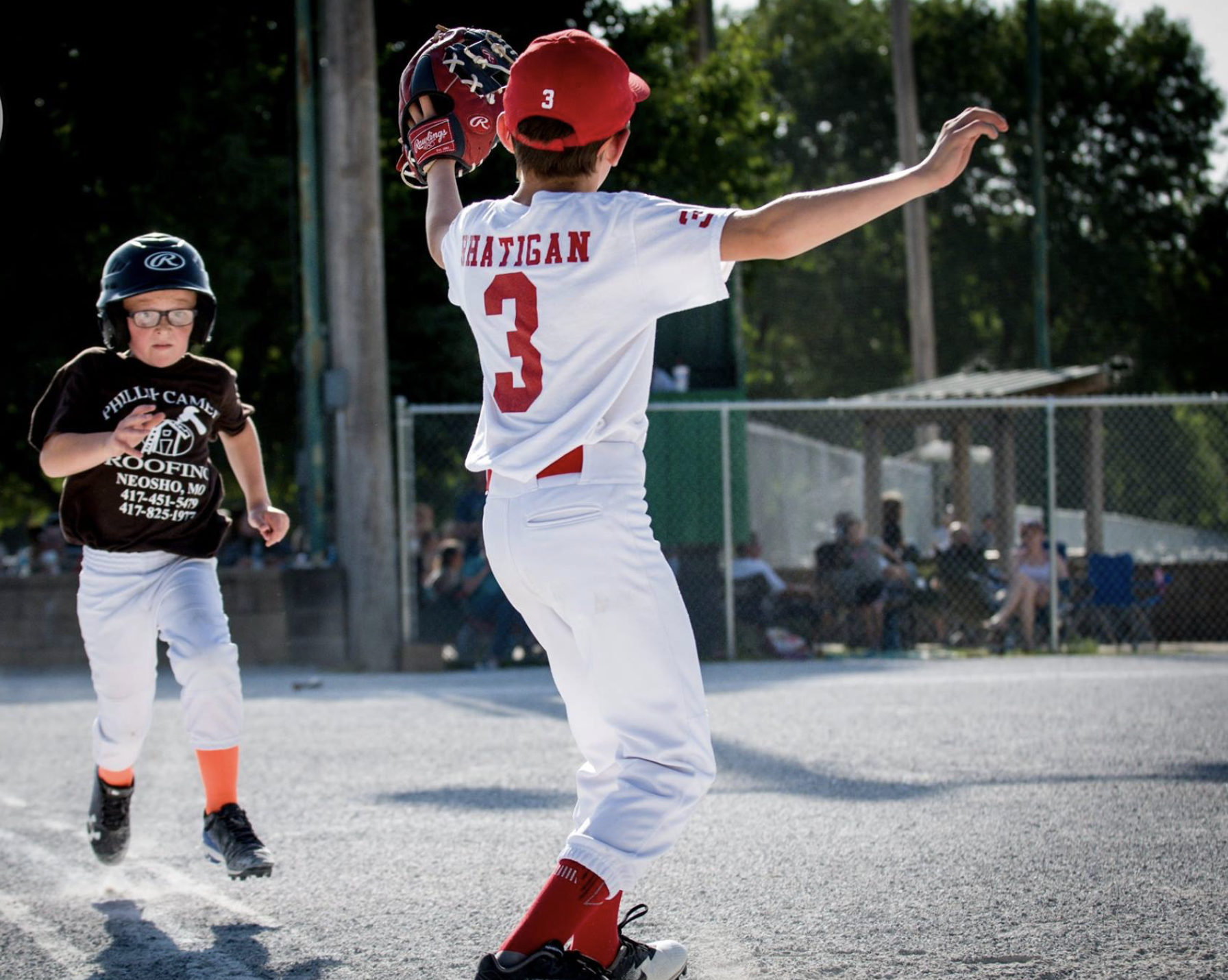 Young baseball players on the field, one wearing a red cap and white uniform with the number 3, and another in a helmet and dark shirt running behind.