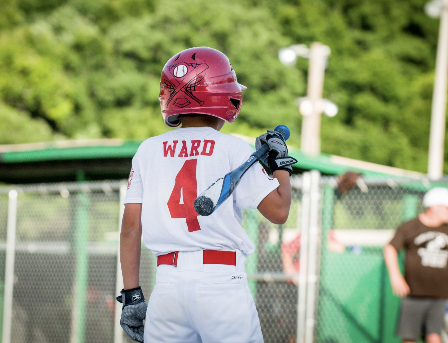 A young baseball player wearing a red helmet, white jersey with number 4 and the name Ward, holding a bat over his shoulder, preparing to bat at a baseball field.