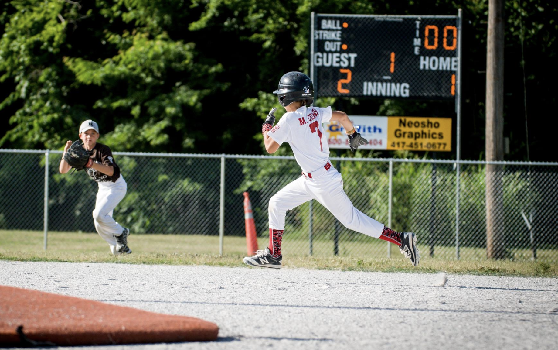 A young baseball player in a white uniform and helmet running towards first base on a baseball field, with another player in black and white waiting with a glove near the base, and a scoreboard in the background.