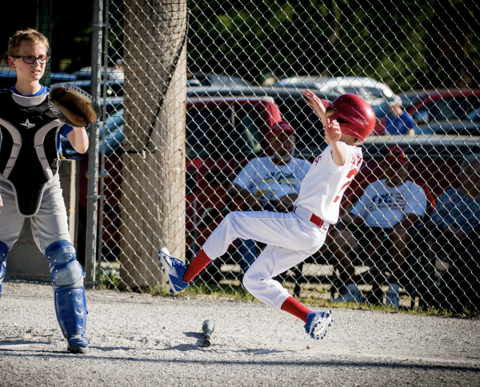 A young baseball player in a white uniform and red helmet slides into home plate while a catcher in protective gear waits nearby.