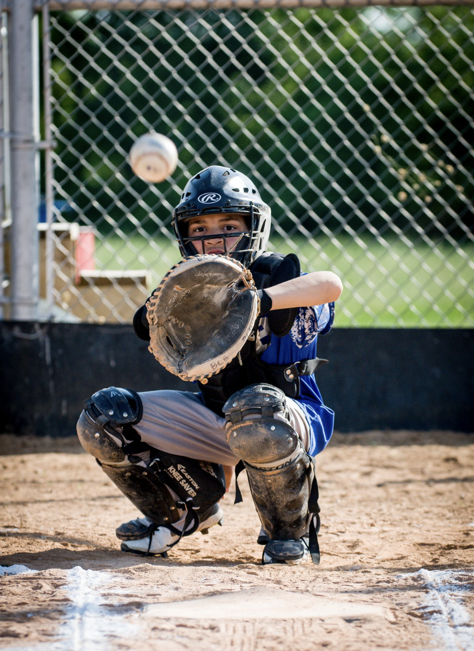 A young baseball catcher crouches behind home plate wearing a helmet, catcher's mitt, and protective gear, ready to catch an incoming baseball.