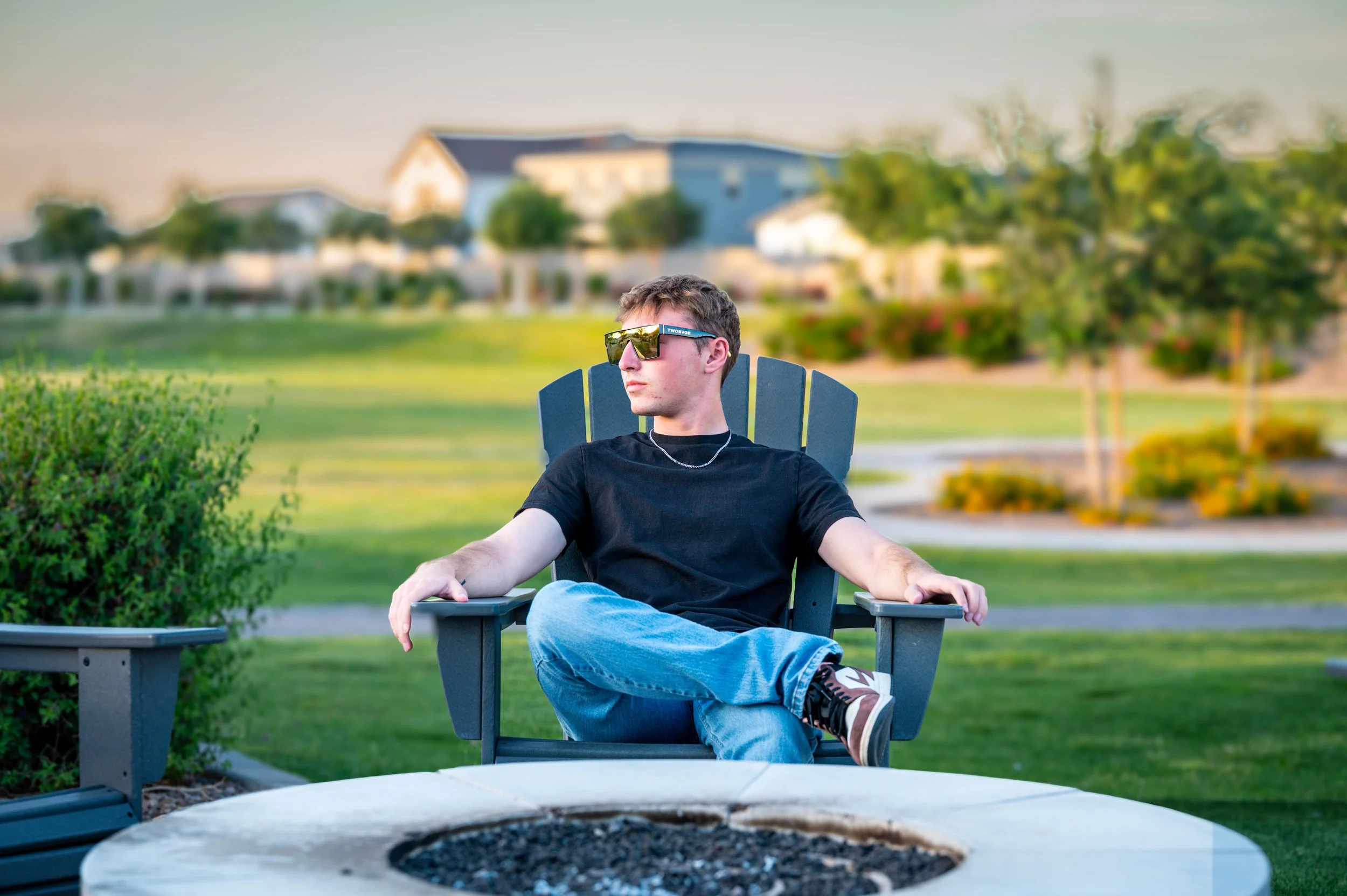 A young man wearing sunglasses, a black t-shirt, and jeans, sitting in a black outdoor chair with his legs crossed, relaxing in a park with green grass, trees, and houses in the background during sunset.