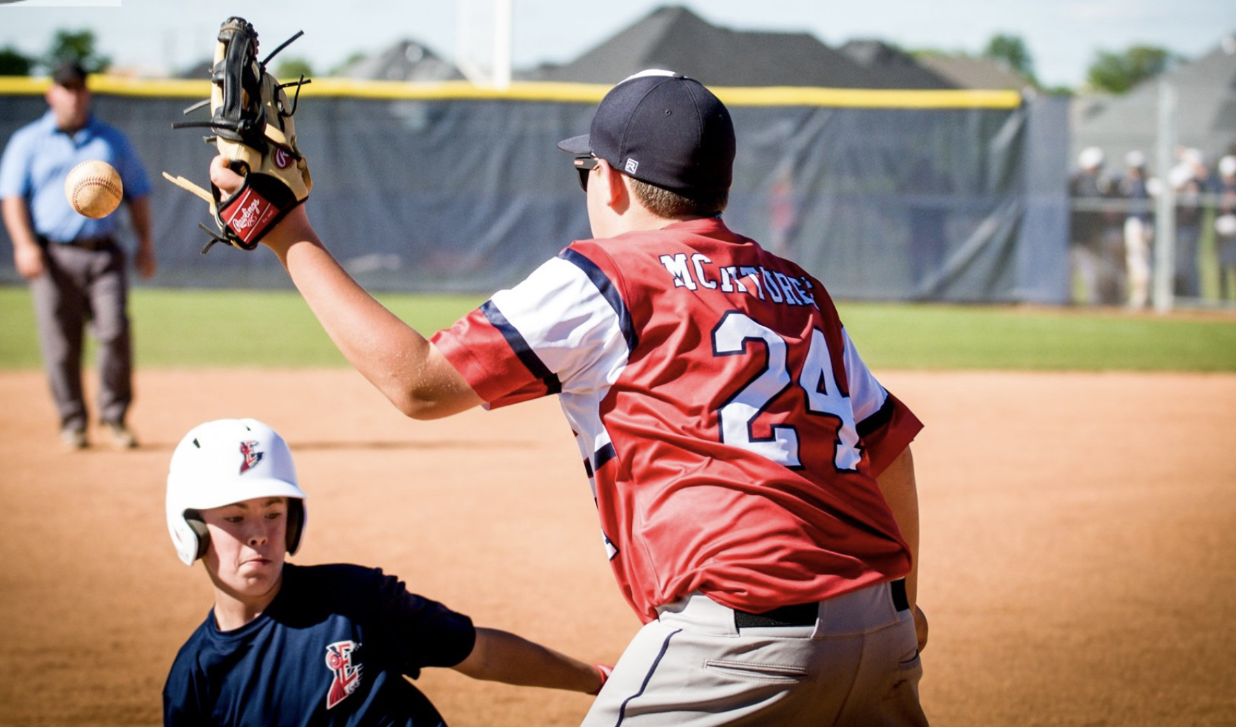A baseball player in a red jersey with the number 24 and the name McFurrey is catching a ball with a glove. A young player in a dark blue uniform and helmet is nearby on the field, with an umpire and other players in the background.