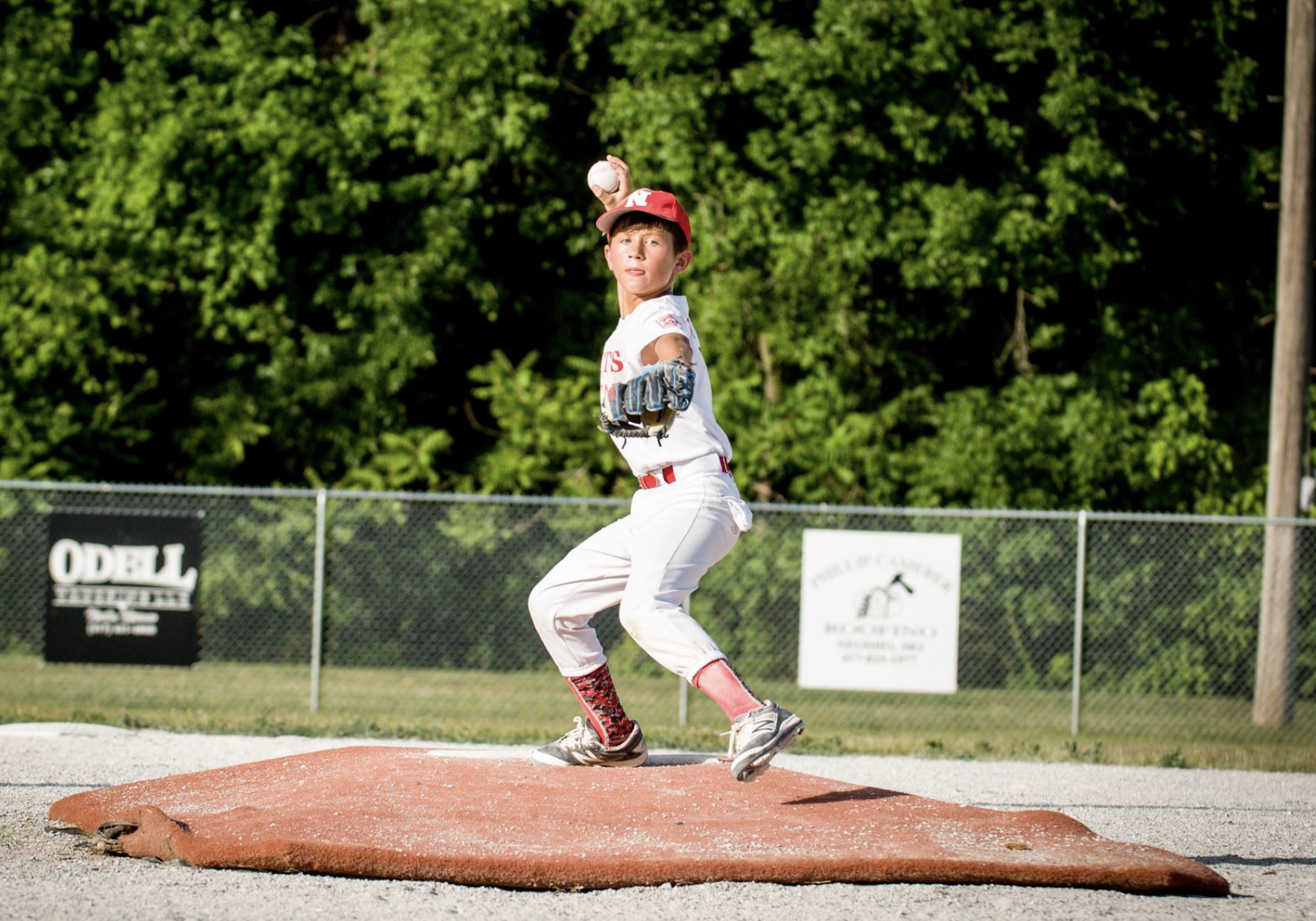 Young baseball player in a white uniform with red accents is pitching on a mound, holding a baseball ready to throw, with a backdrop of green trees and a chain-link fence.