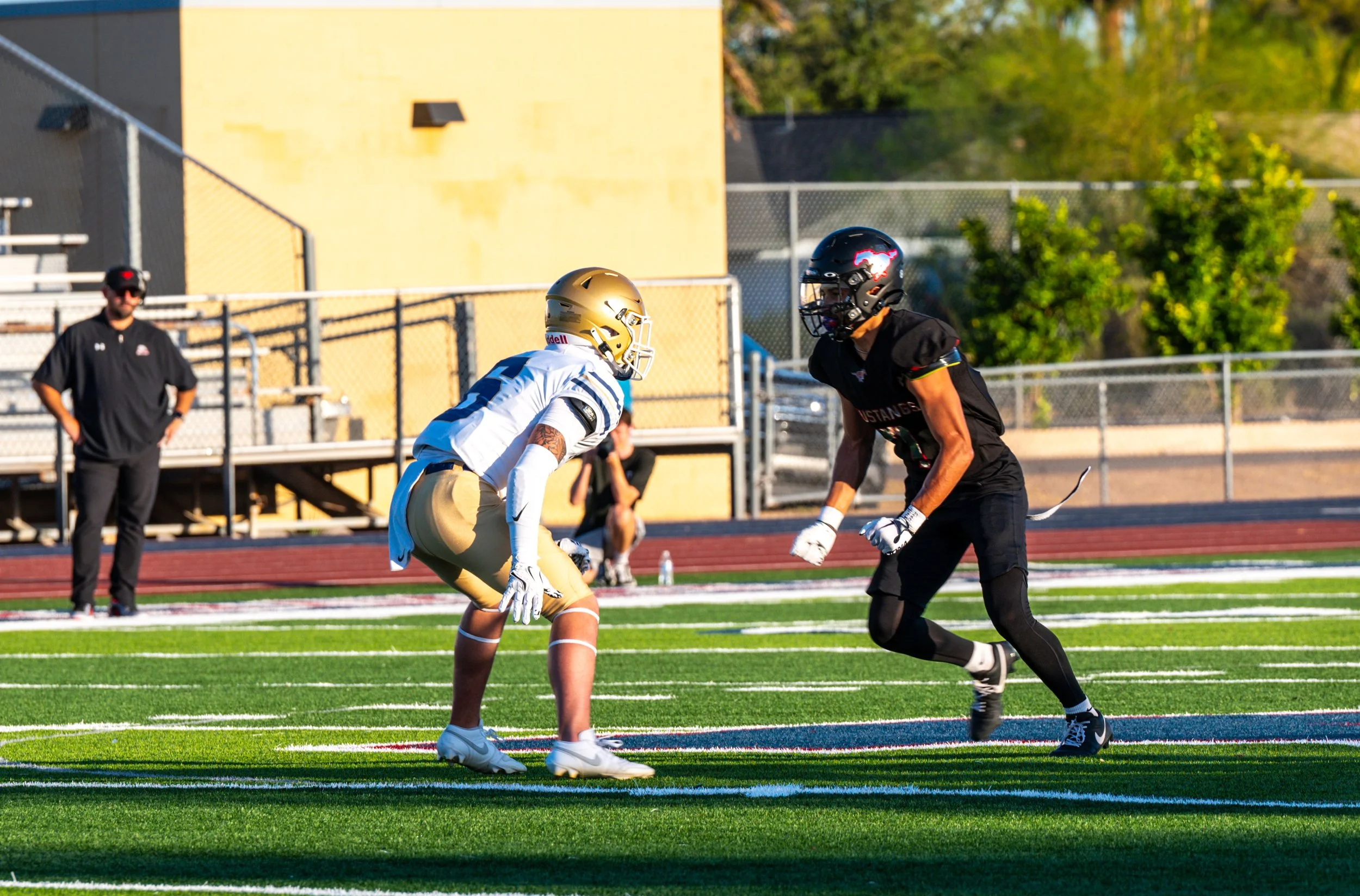 Two football players in helmets and uniforms facing each other on a football field, about to engage in a play, with a coach and trees in the background.