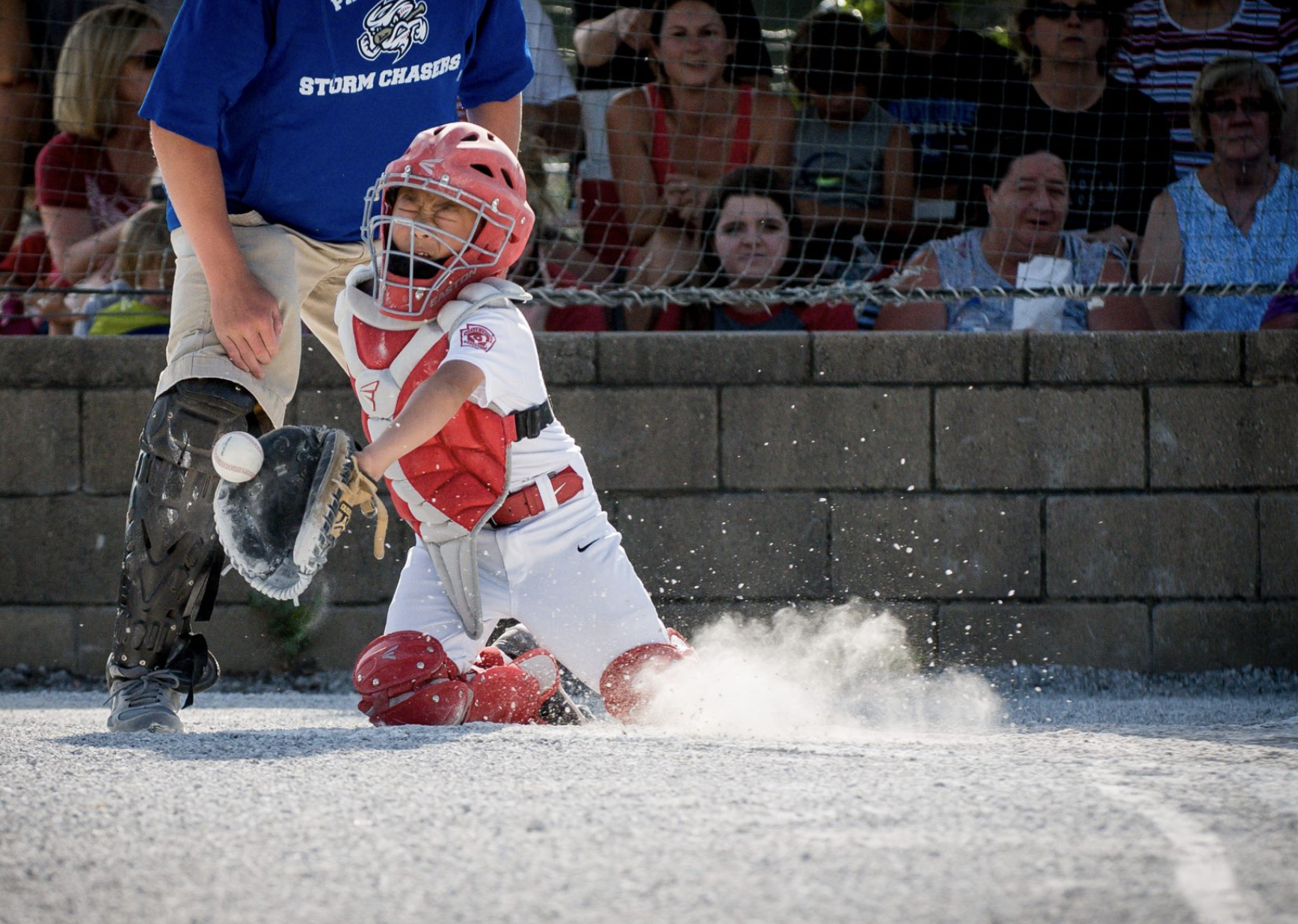 A young baseball catcher in a red and white uniform is on his knees, reaching for a ball on the ground, with an umpire nearby and spectators watching behind a chain-link fence.