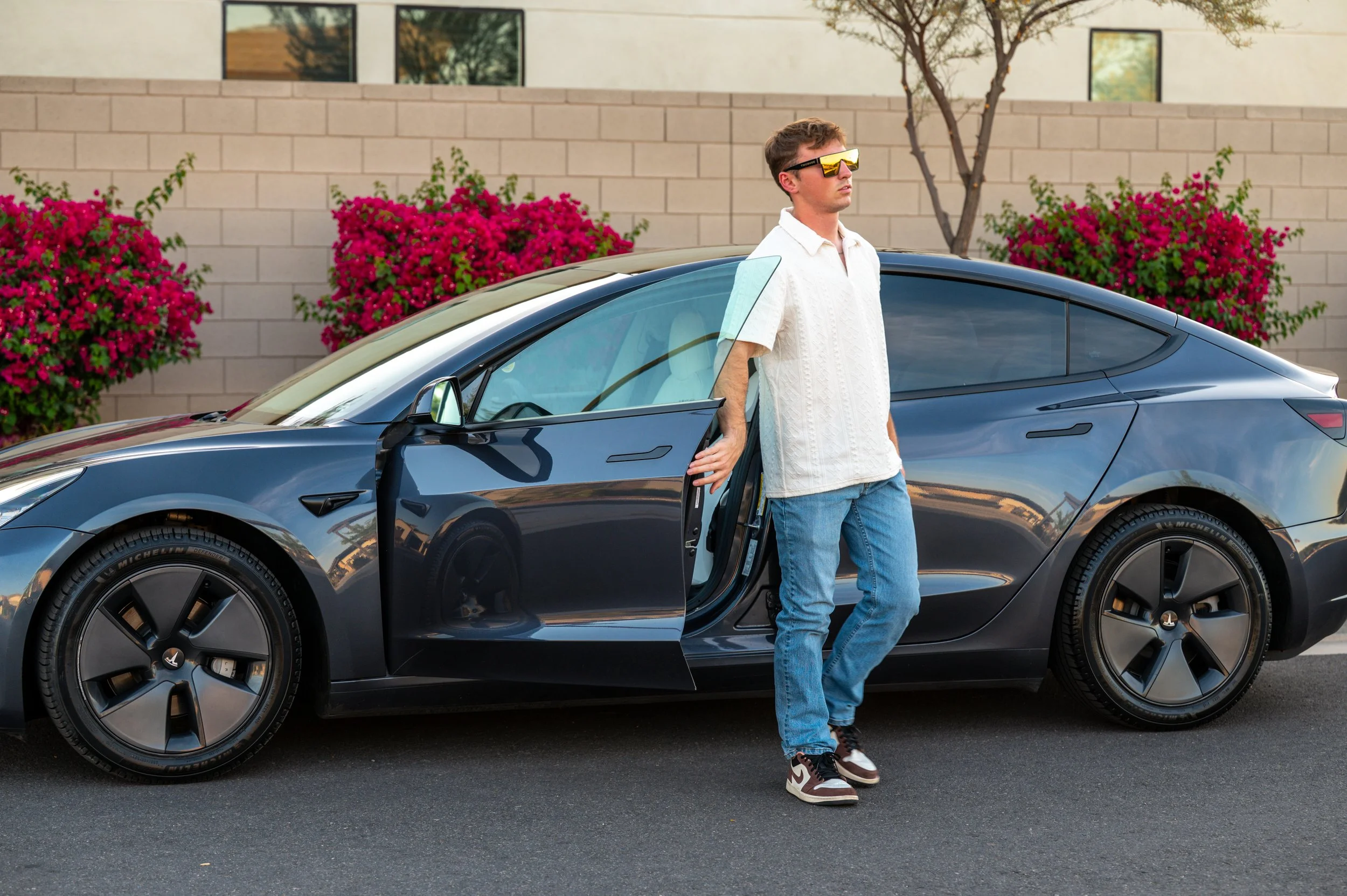 Young man in white shirt and jeans standing outside next to a black Tesla car with the door open, wearing futuristic sunglasses. There are pink flowering bushes and a beige building in the background.